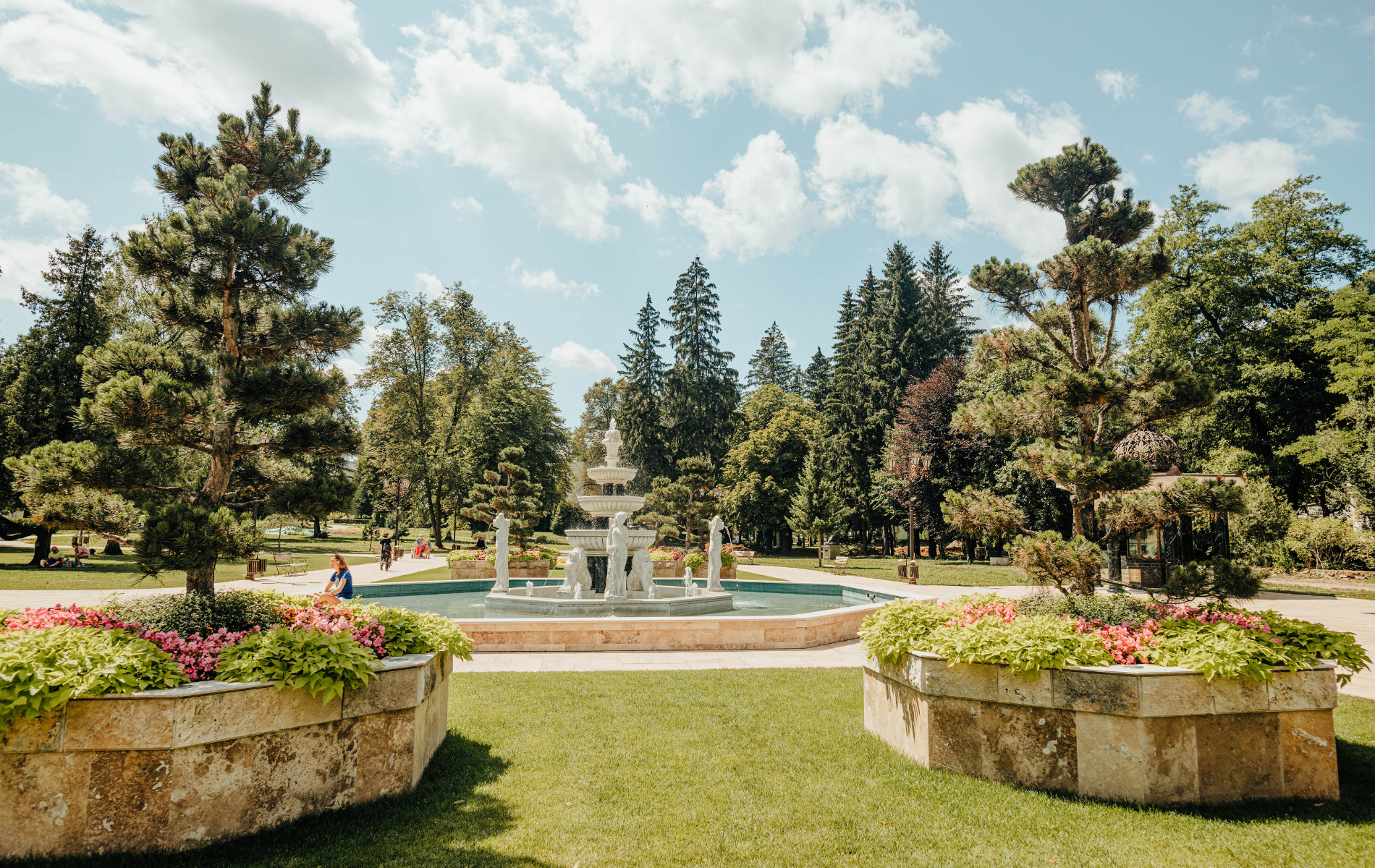 Formal garden with a fountain, trees, flowers, and a partly cloudy sky.
