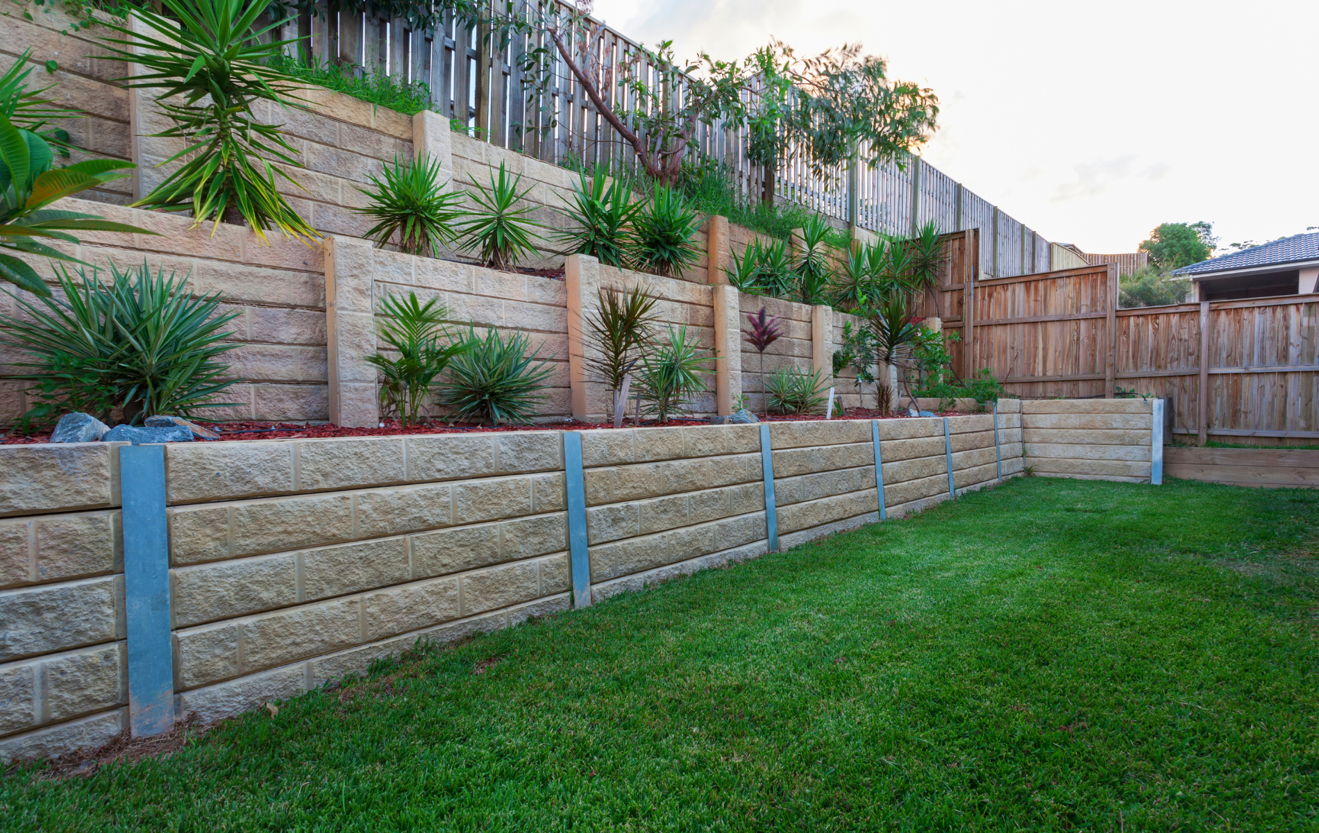 Retaining wall with tiered landscaping, green grass, and a wooden fence in a yard.