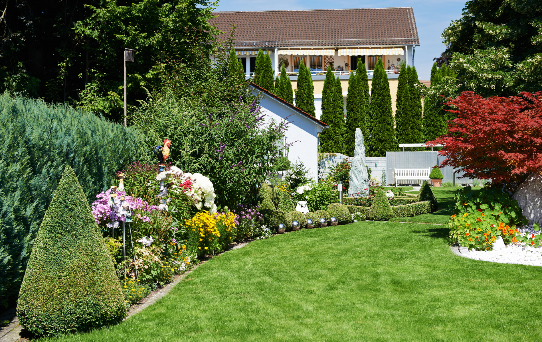 Well-manicured garden with flowers, trees, and a small white structure, in front of a light-colored building.