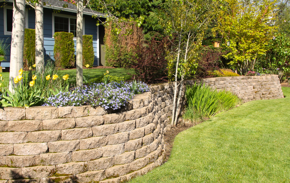 Stone retaining wall with tiered flower beds, grass lawn, and house in background.