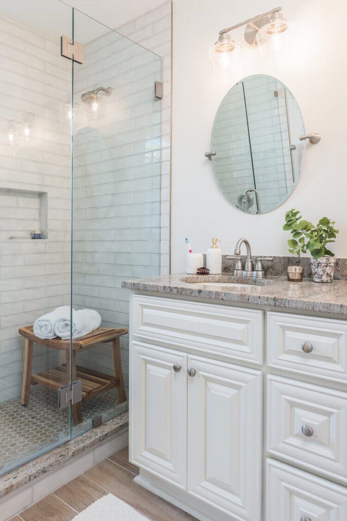 Bright bathroom with white cabinets, granite countertop, glass shower, and oval mirror.