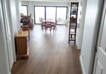 Hallway leading to a dining area with a table, chairs, and a balcony view. Brown wooden floors, light walls.