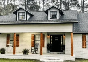White brick house with brown shutters and porch posts, black door, and two dormers with black trim.