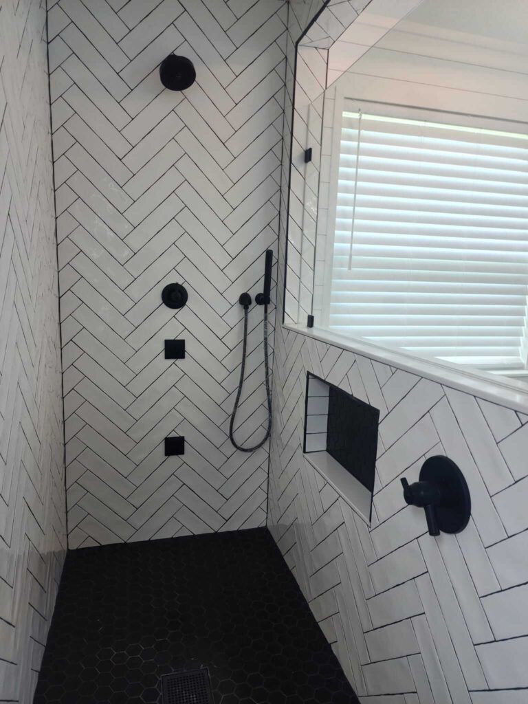 A modern shower with white herringbone tiles, black fixtures, and dark floor.