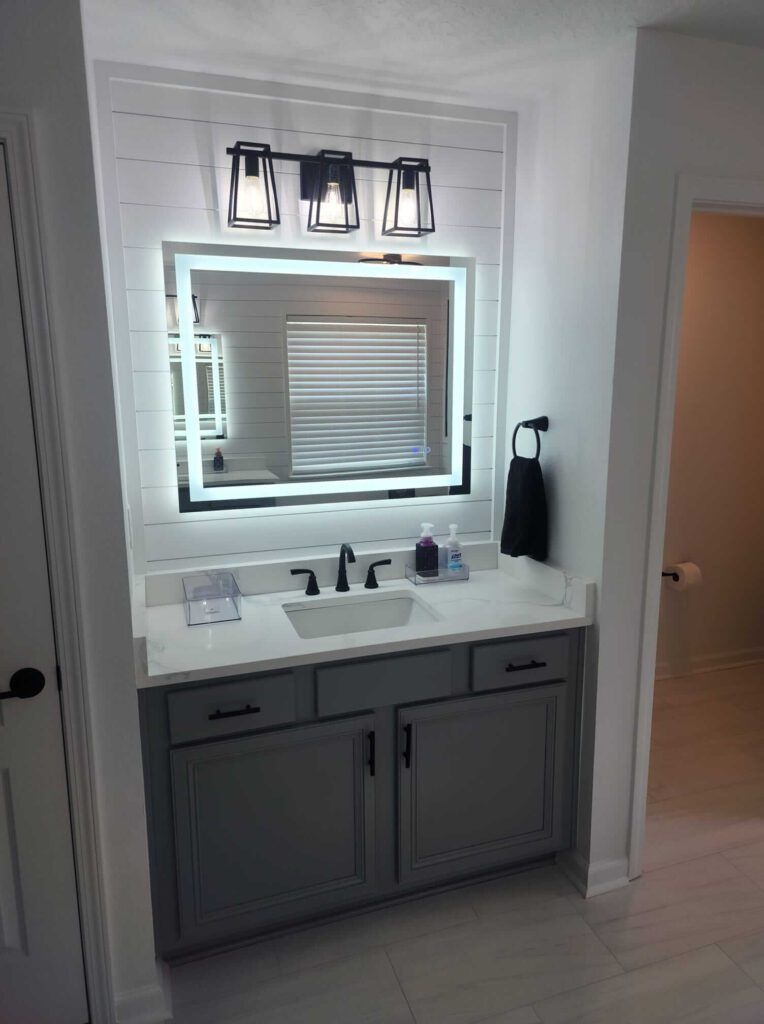 Bathroom with gray vanity, lit mirror, black fixtures, and three-light fixture. White wall.