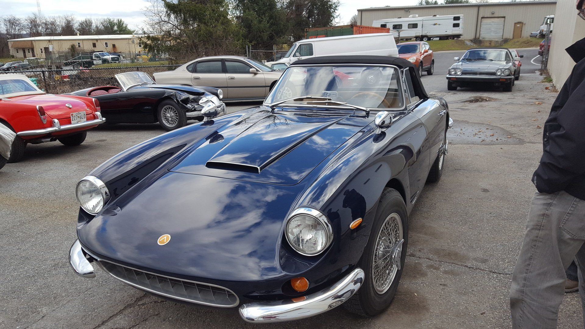 Dark blue vintage convertible parked in a lot, other classic cars and a building in the background.