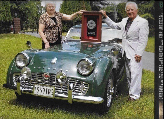 A couple poses with a vintage green Triumph TR3 sports car, holding an award on the hood.