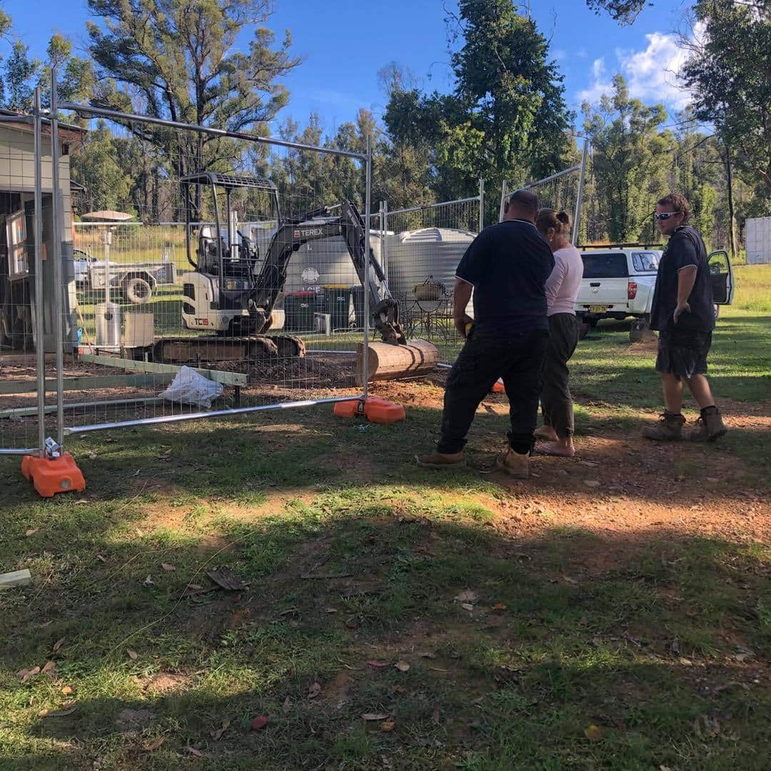 A Group of People Are Standing in A Grassy Field in Front of An Excavator — Alcando Renovations & Maintenance In Taree, NSW