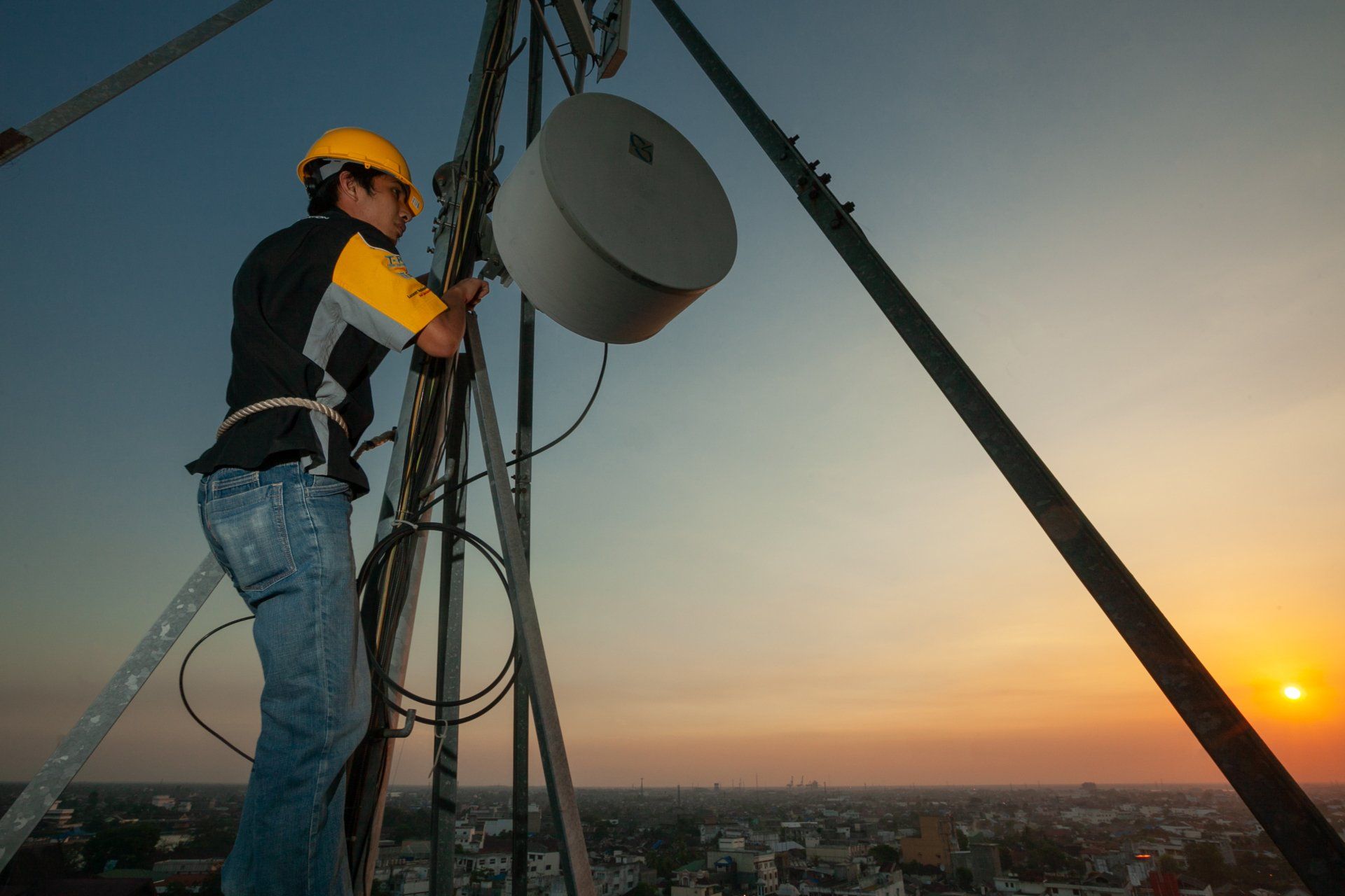 A man in a hard hat is working on a tower at sunset