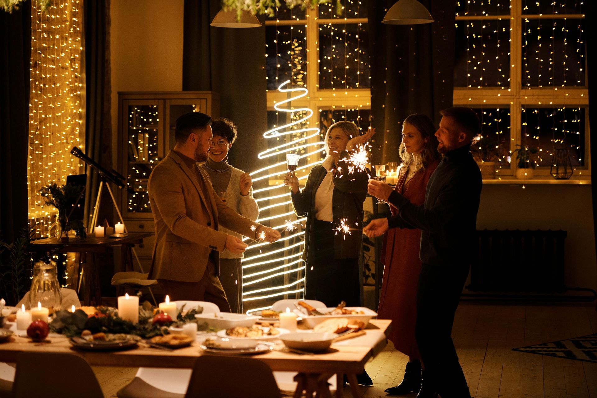 People at a holiday dinner party, holding sparklers near a decorated Christmas tree and candles.