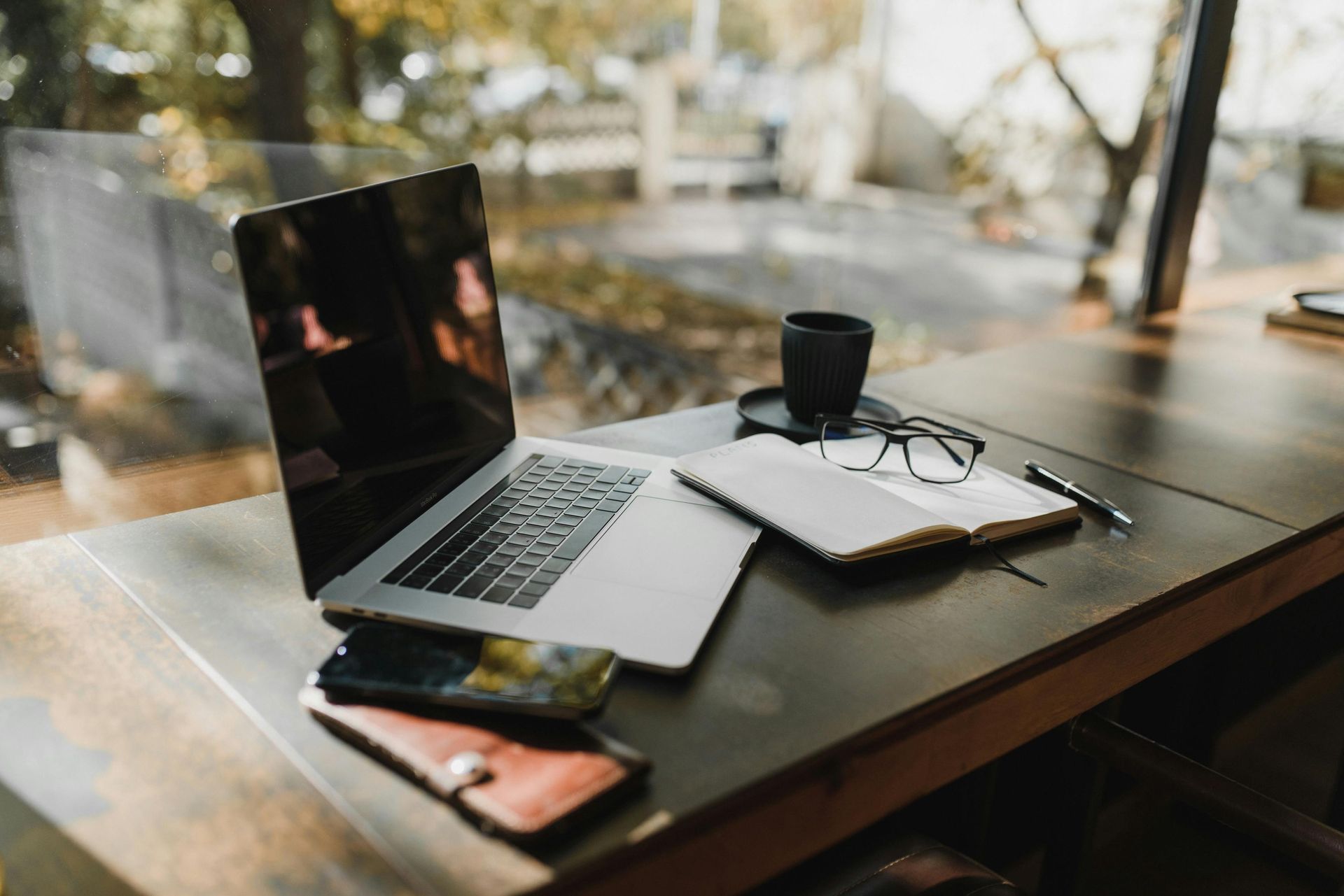 Laptop, notebook, glasses, phone, and coffee cup on a wooden table near a window.