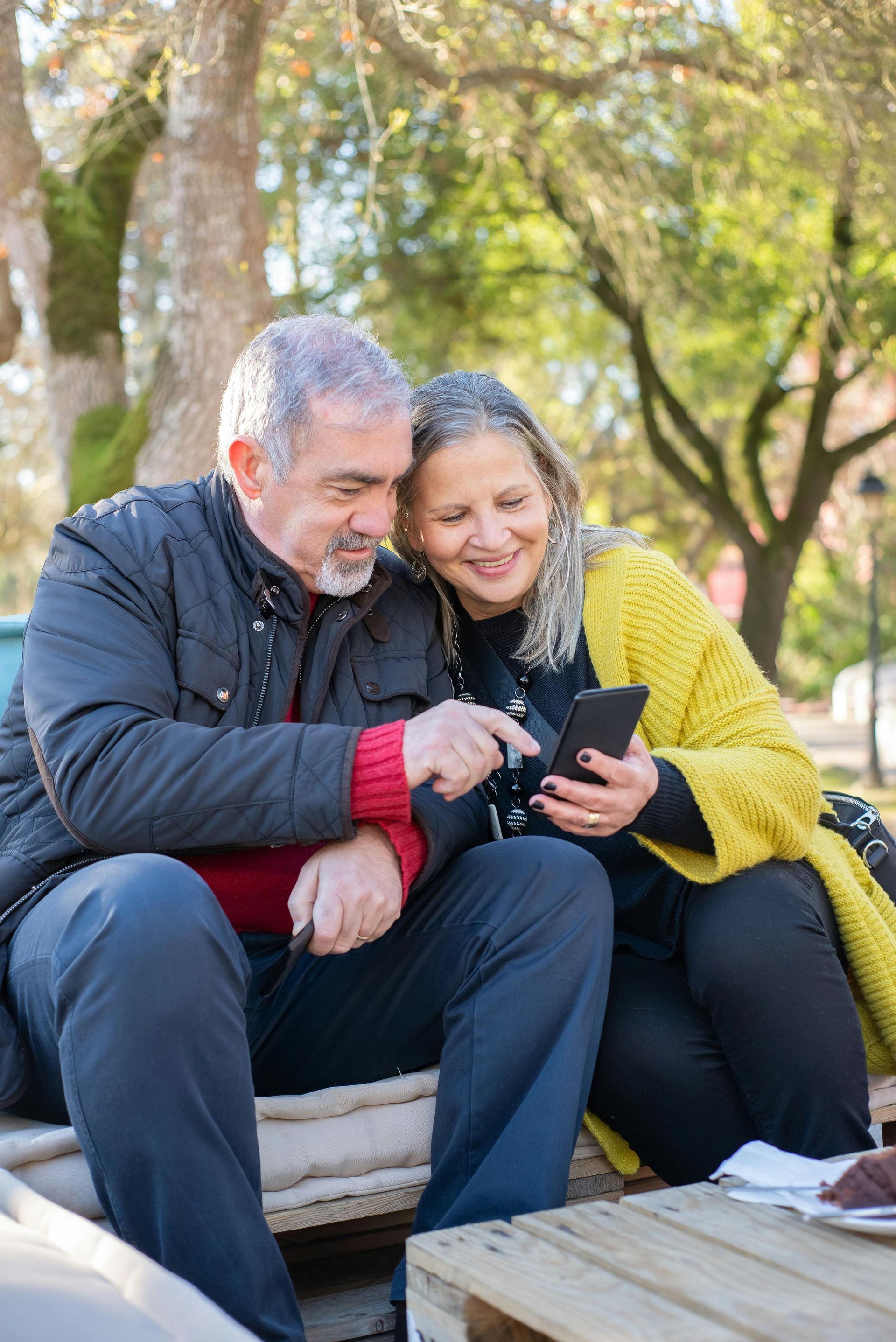 Older couple smiles while looking at a phone together outdoors, man points at the screen.