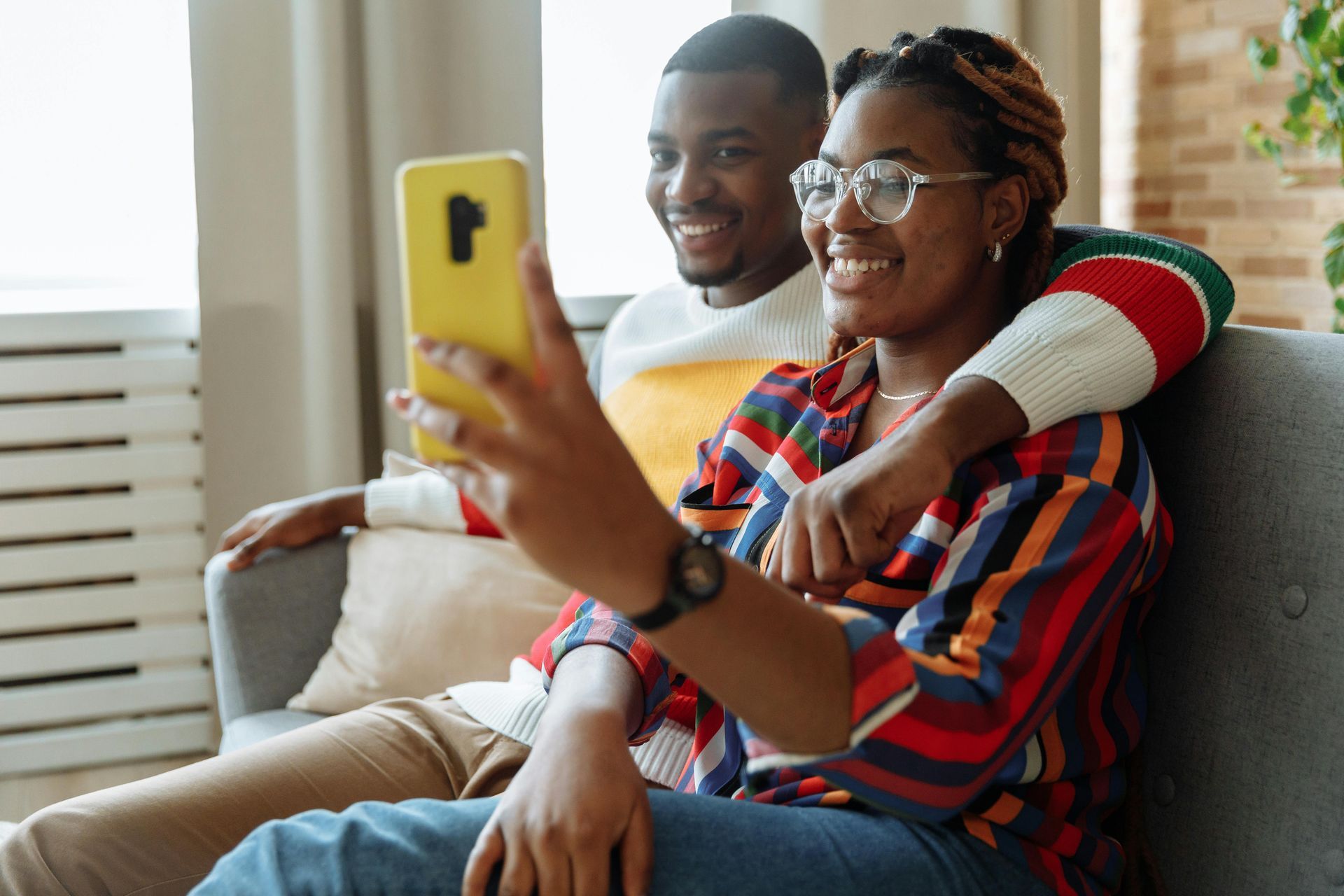 Couple sitting on a couch, woman holding yellow phone and smiling, arm around her, taking selfie.