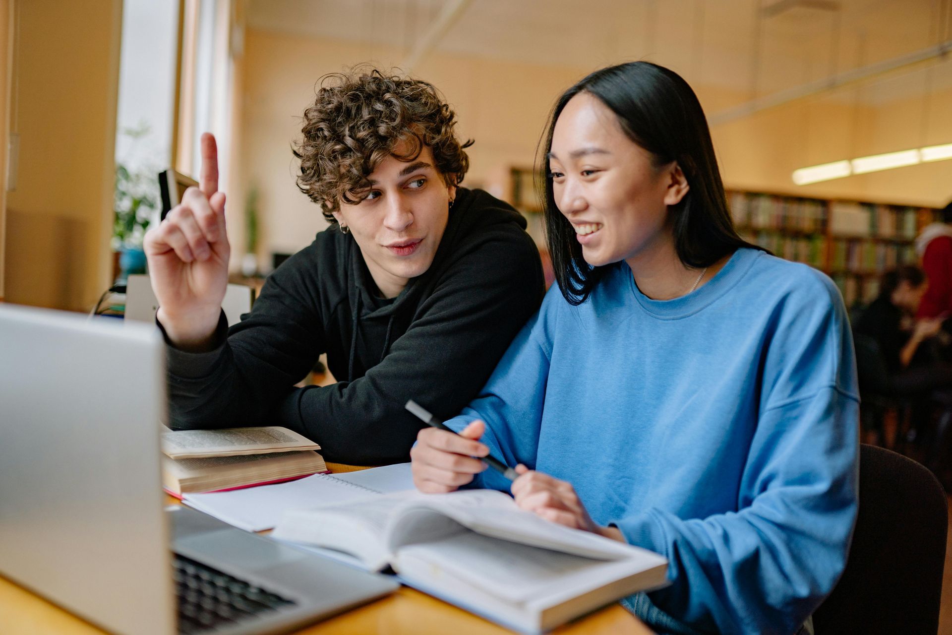 Two students studying together at a library, using a laptop and books.