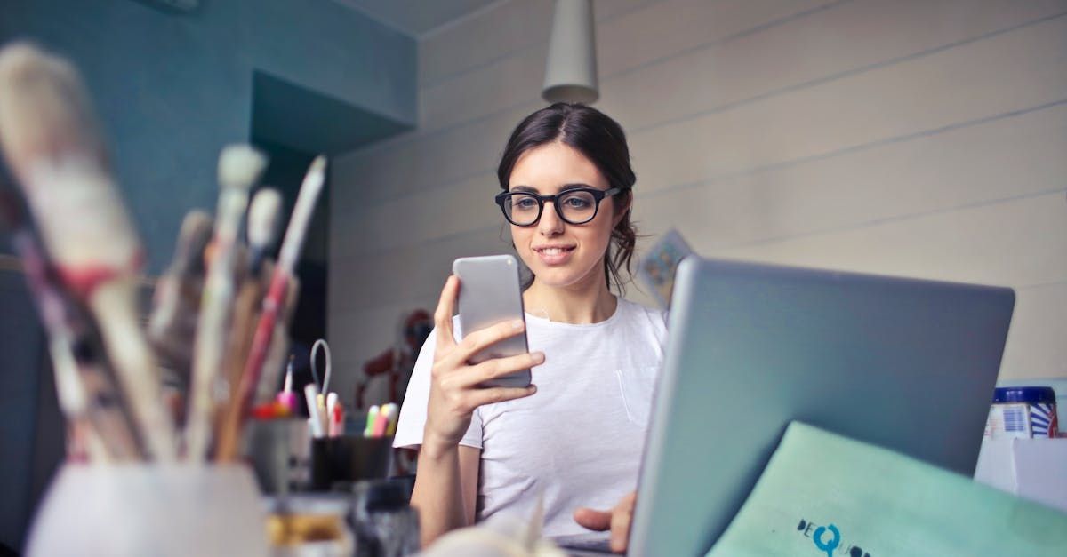 A woman is sitting at a desk using a laptop and a cell phone.