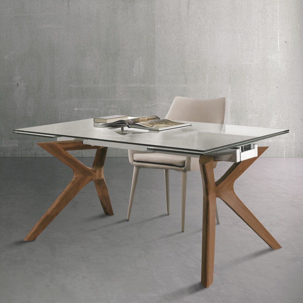 Tavolo da pranzo con piano in vetro e gambe a stella in legno; dietro c'è una sedia.