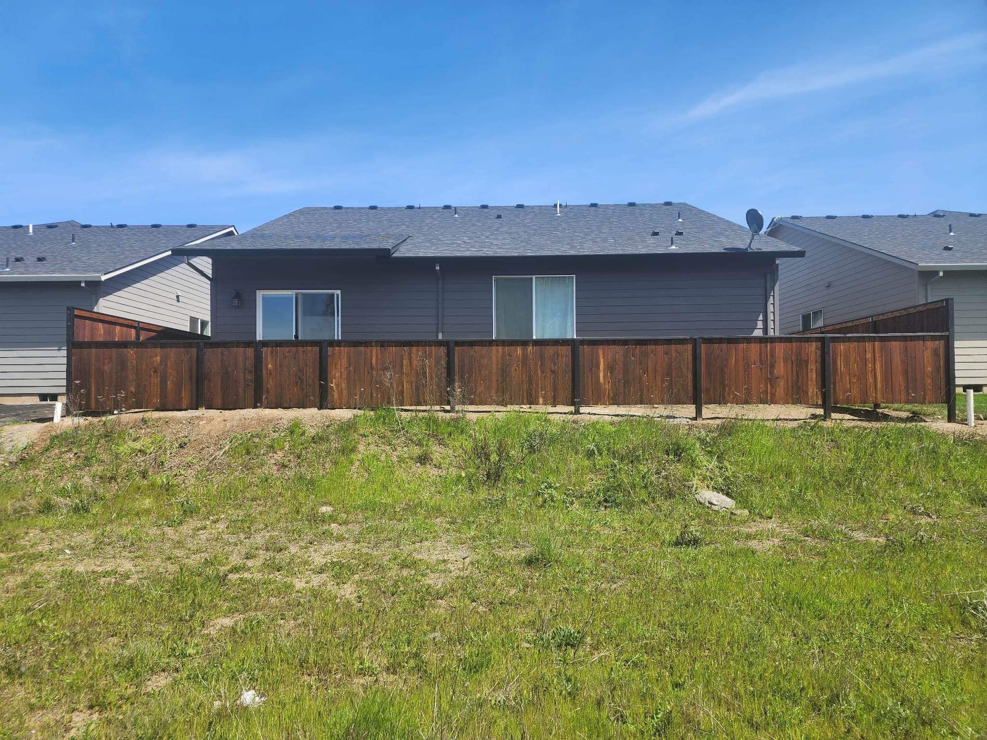 Suburban house with a fenced backyard and grassy field in front under a clear sky