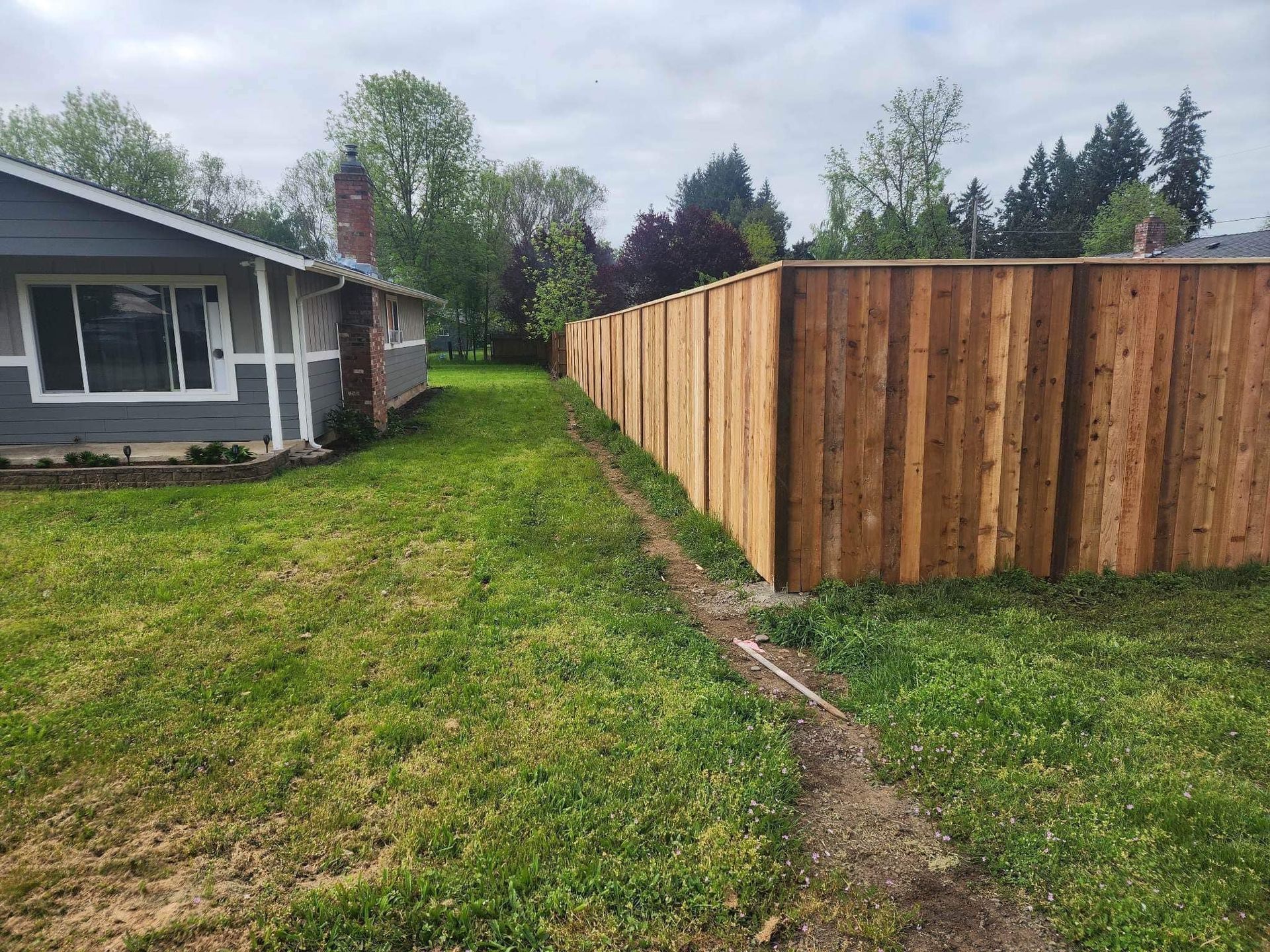 Side yard with green lawn beside a house and a long wooden fence under a cloudy sky