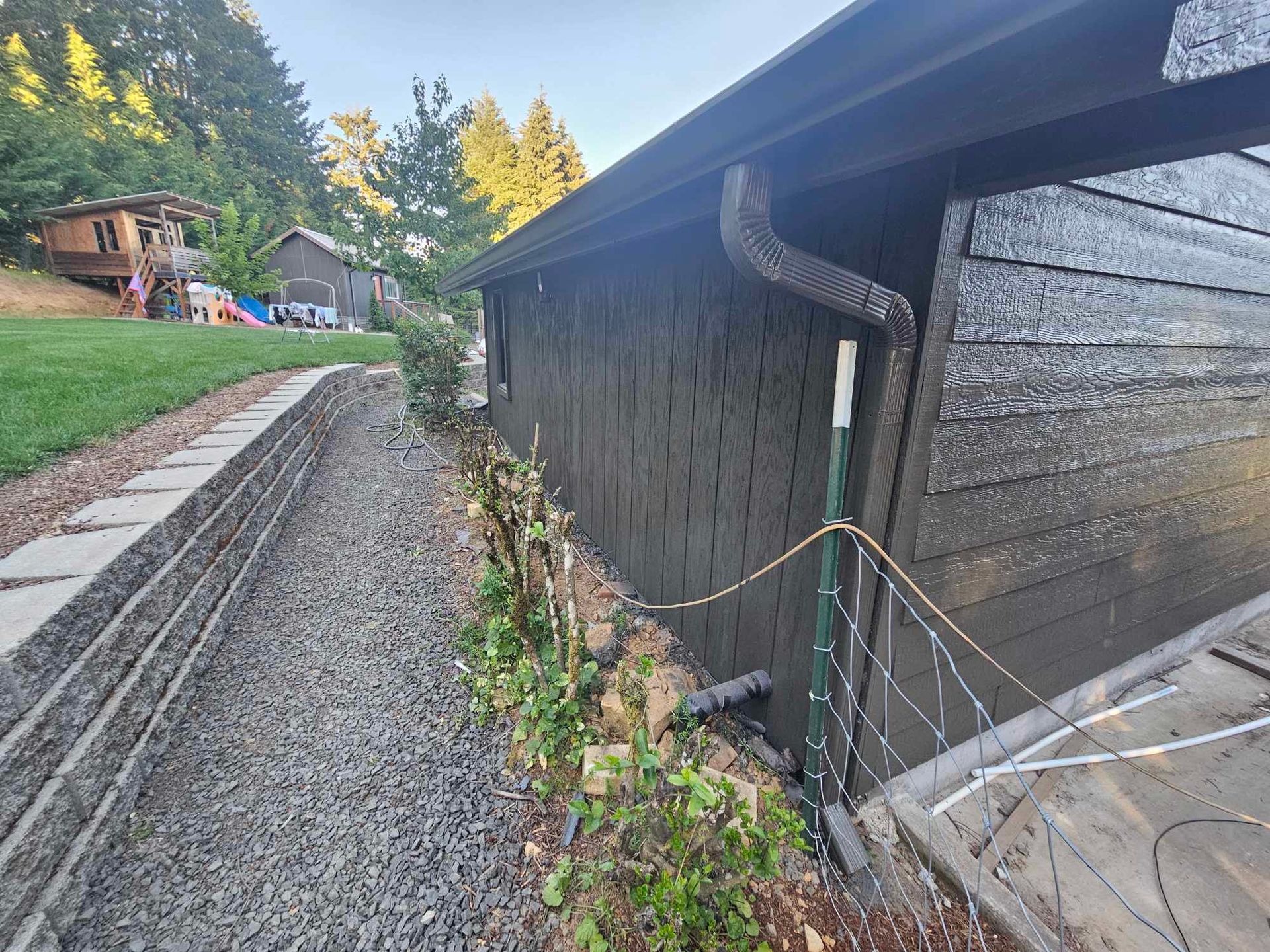 Gravel path beside a black building with a small garden and hillside homes in the background