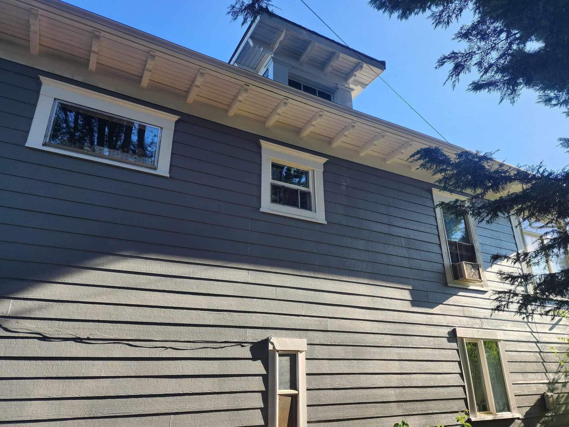 Gray house exterior with small windows and a sunny blue sky above, partly framed by tree branches
