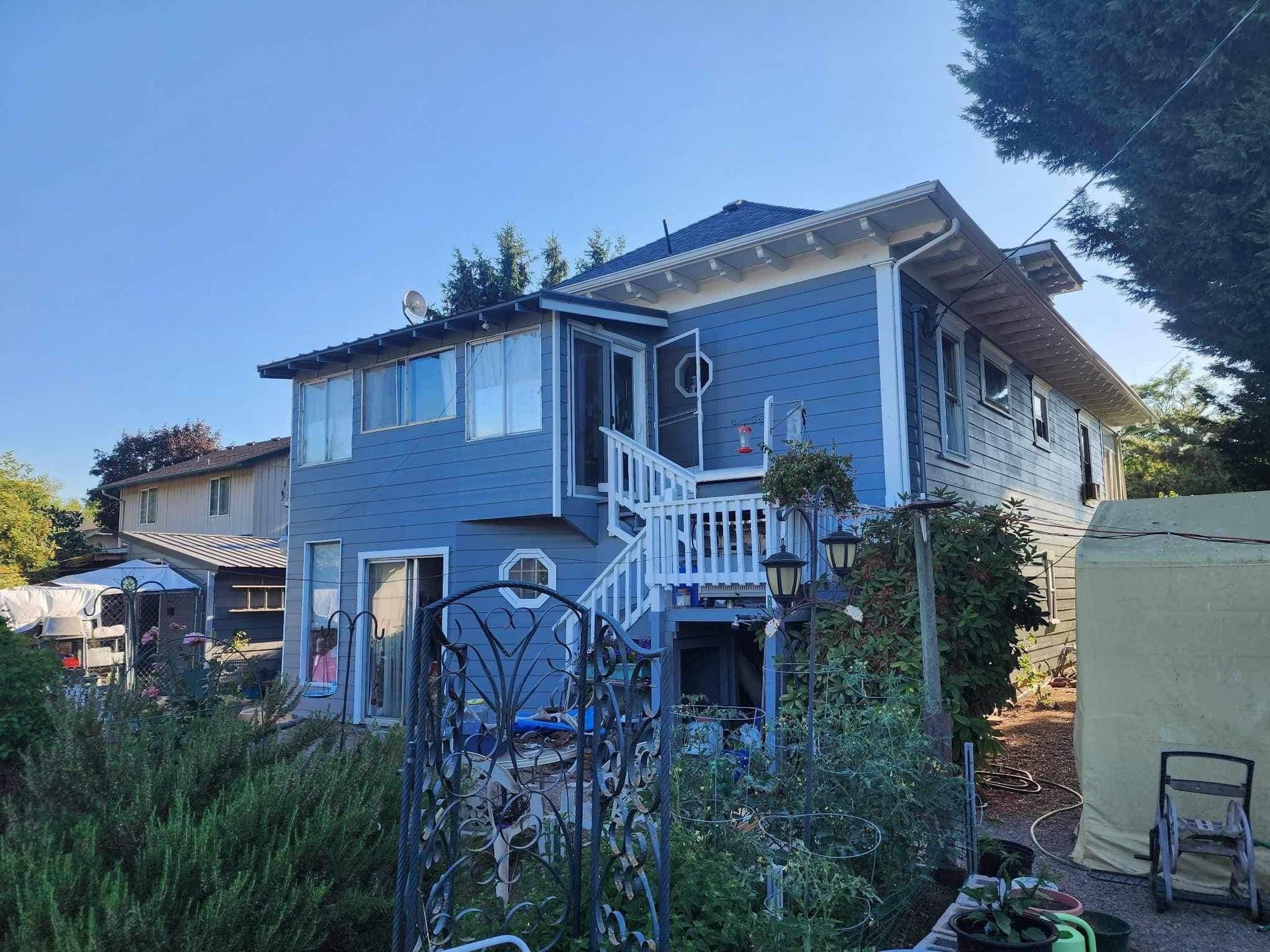 Blue two-story house with white trim, front porch, and overgrown yard behind a metal gate.