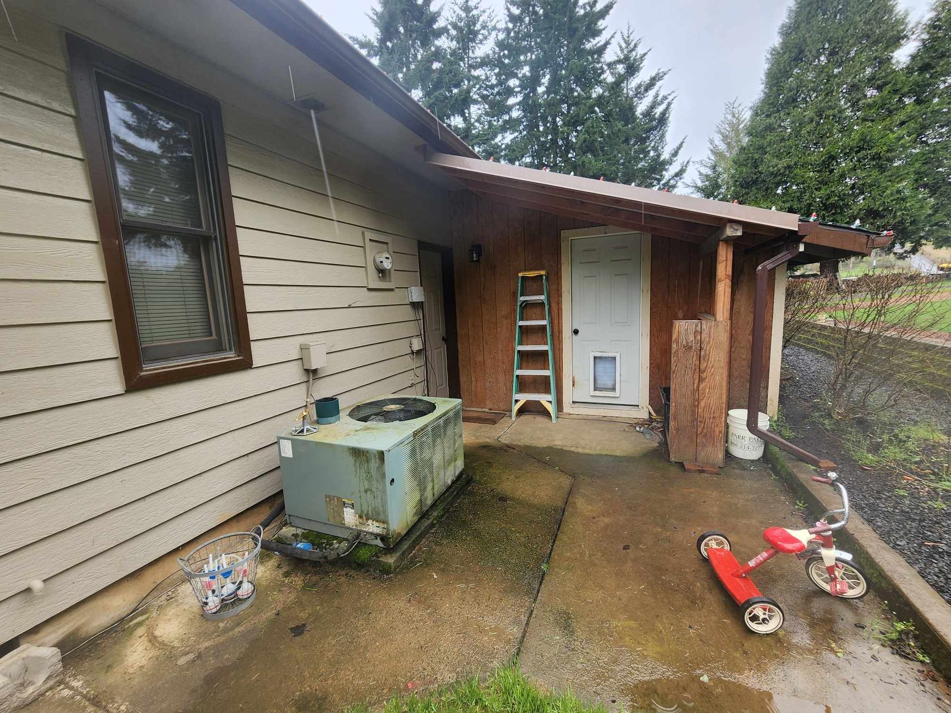 Side yard patio with a white door, ladder, hose, and scattered tools beside a beige house
