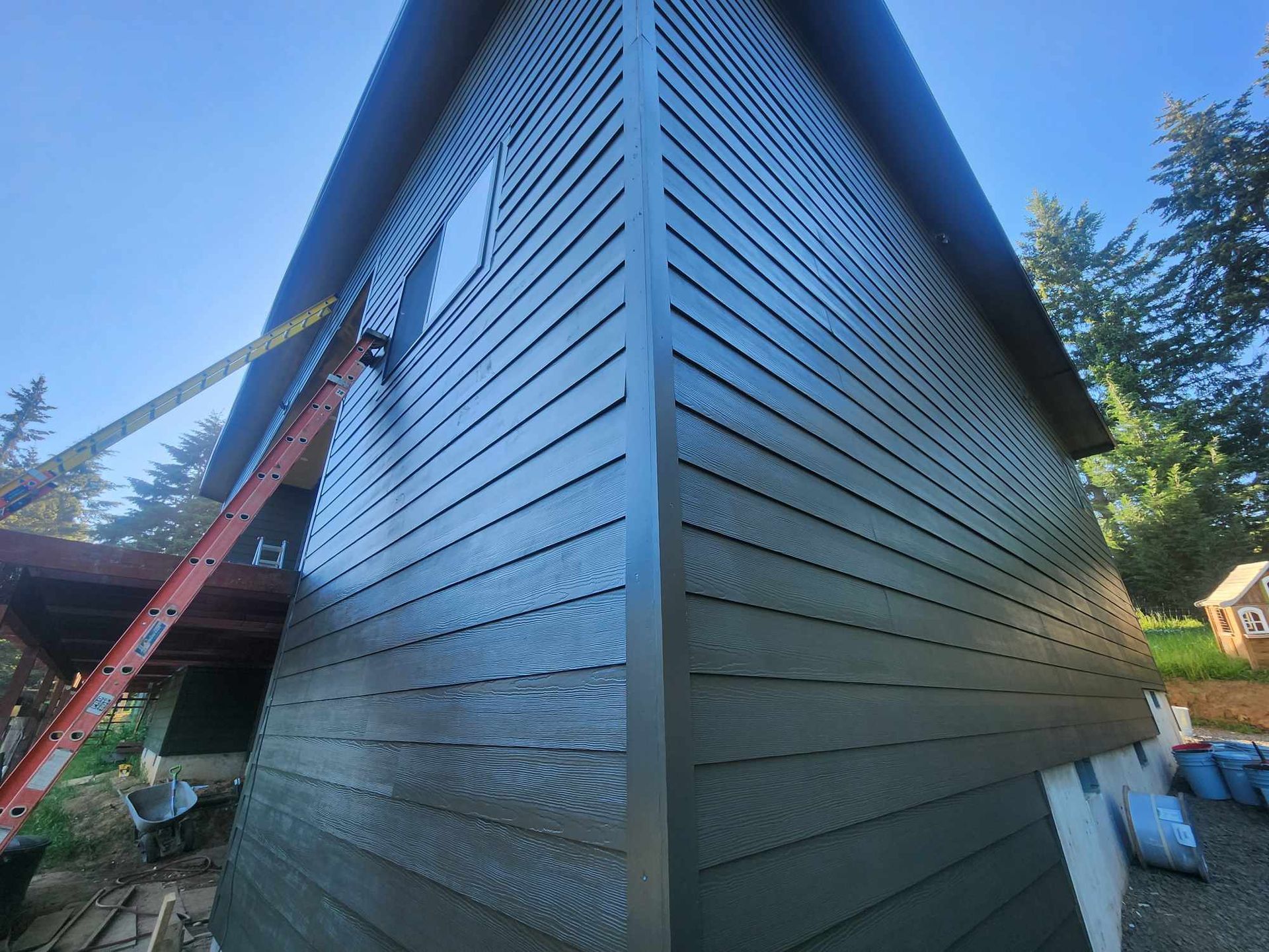 Exterior of a modern house with dark blue siding, a red ladder, and trees under a clear sky