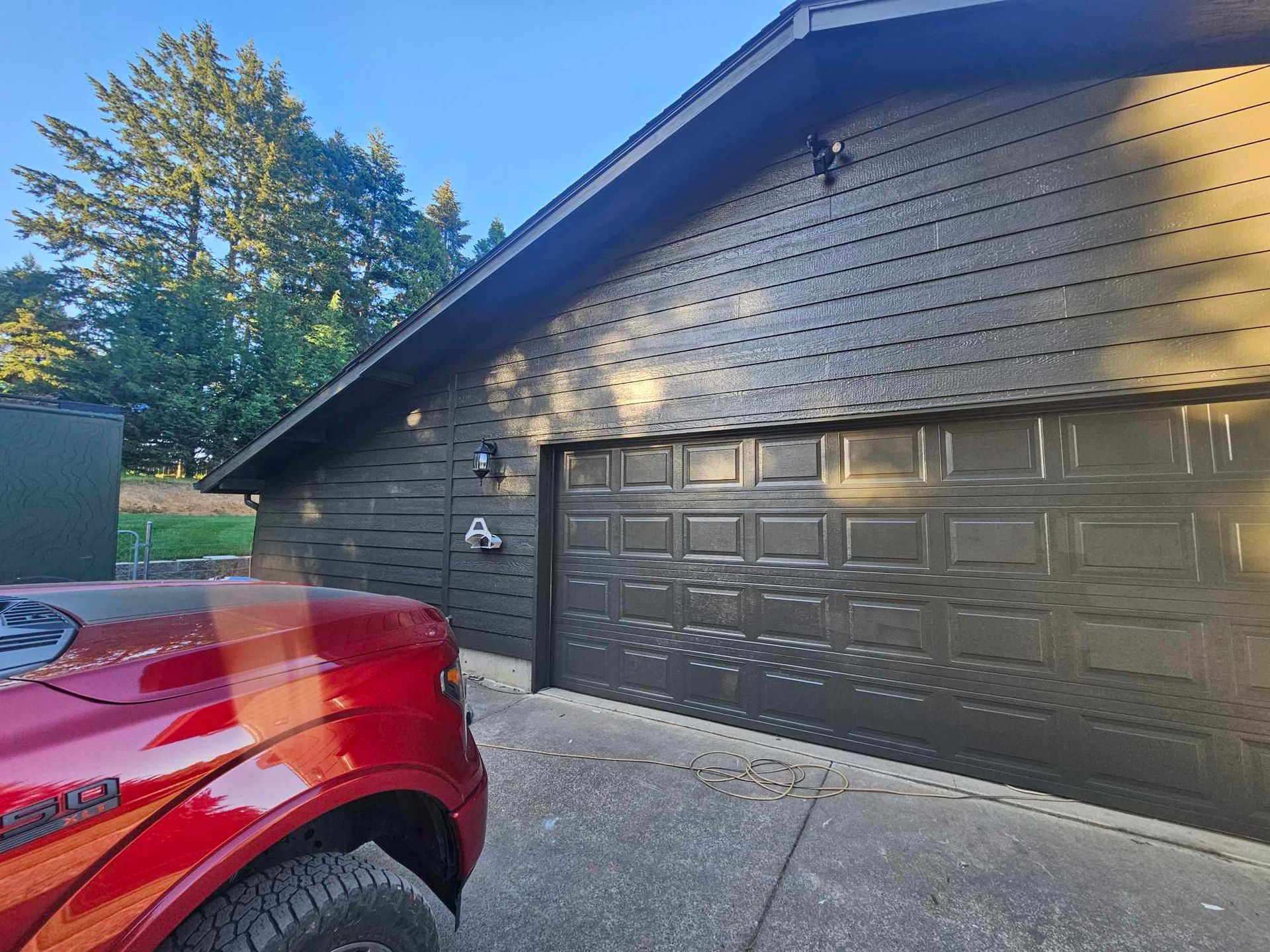 Red pickup truck beside a dark garage with a textured black exterior and sunlit trees nearby