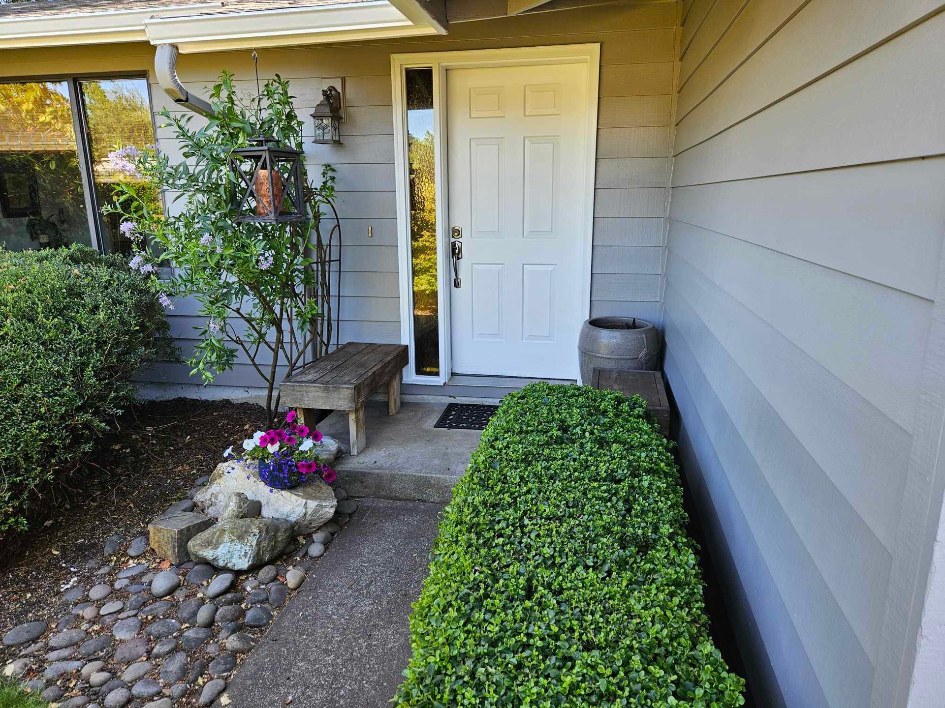 Front porch with white door, potted flowers, stone edging, and trimmed green shrubs beside beige siding.