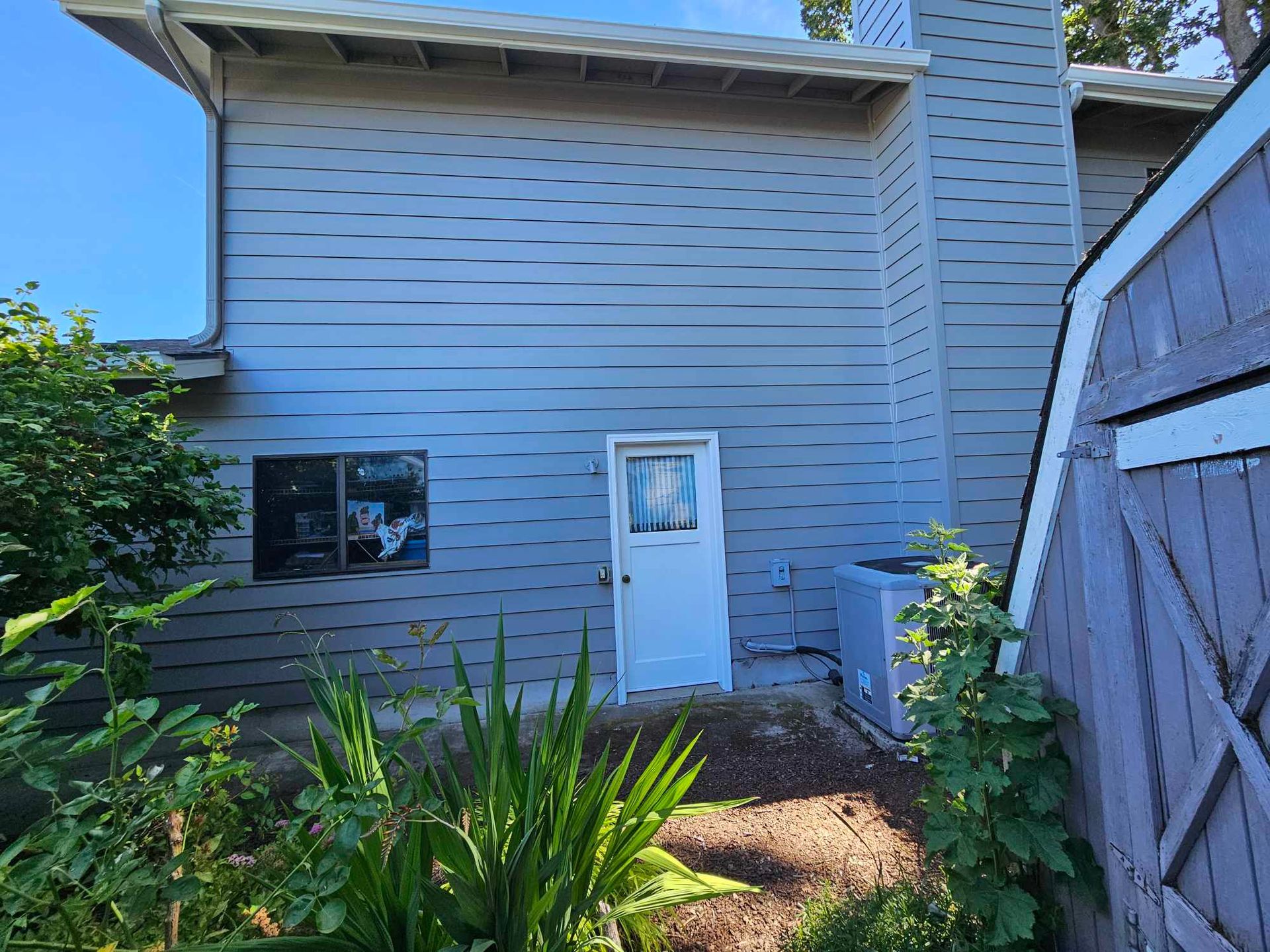 Backyard view of a white house with a door, window, and garden plants beside a wooden fence