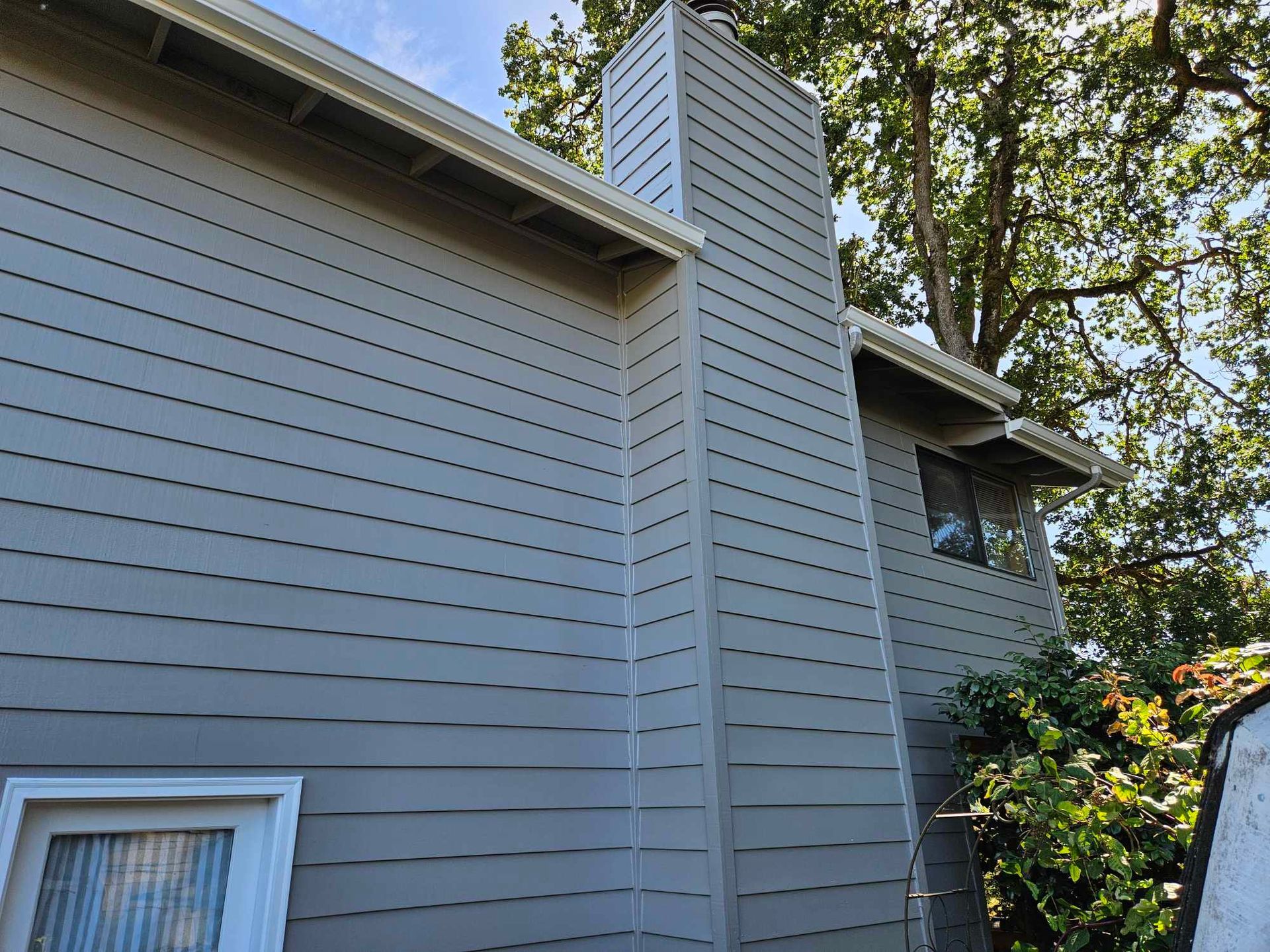 Gray house exterior with white-trimmed windows and a central chimney, framed by trees and blue sky