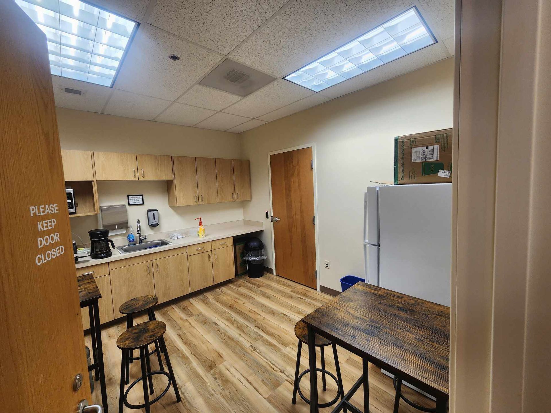 Small break room with cabinets, sink, tables, stools, and a refrigerator under bright ceiling lights.