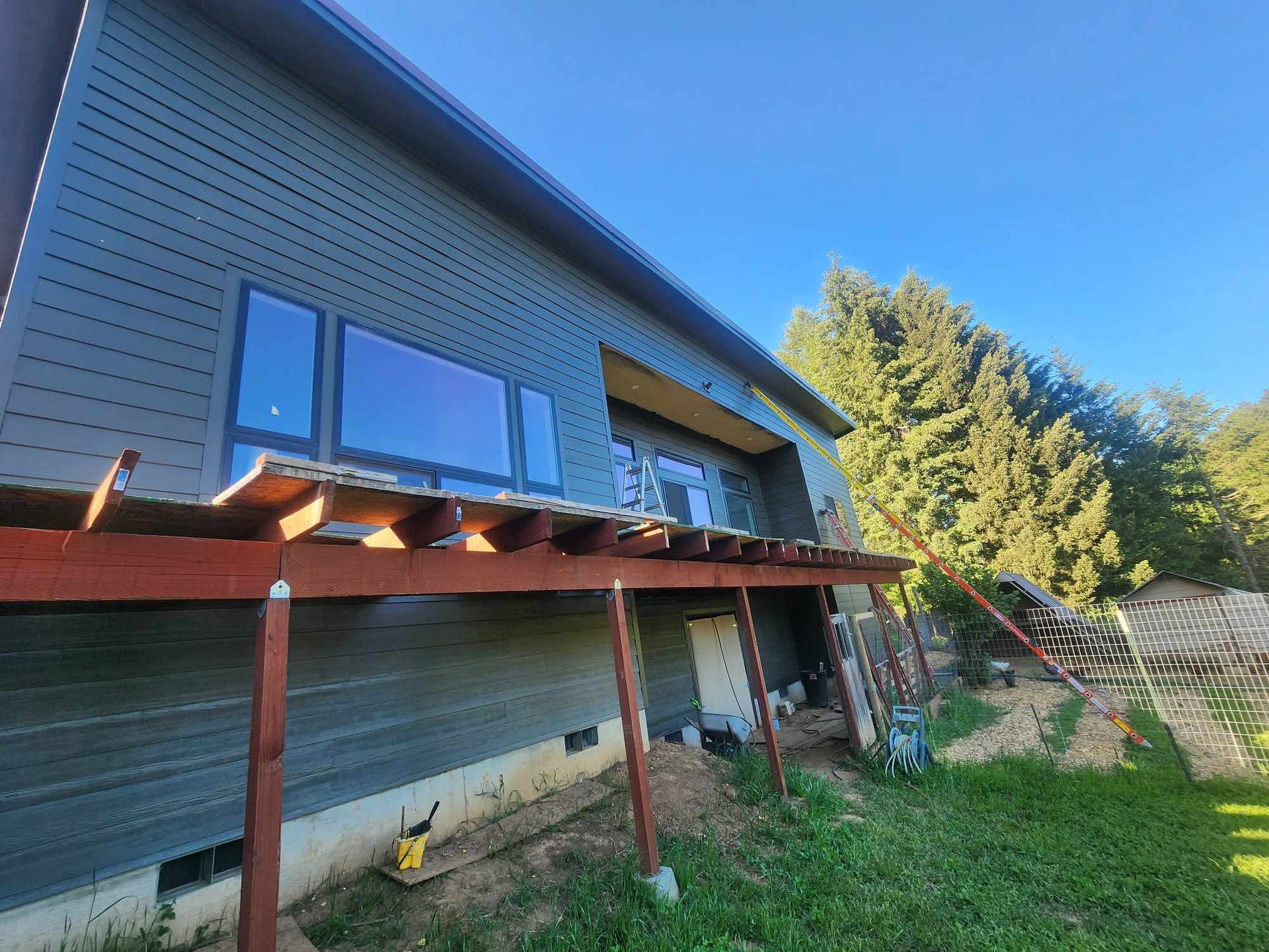 Rusty backyard deck beside a gray house under a blue sky, with grass and trees nearby