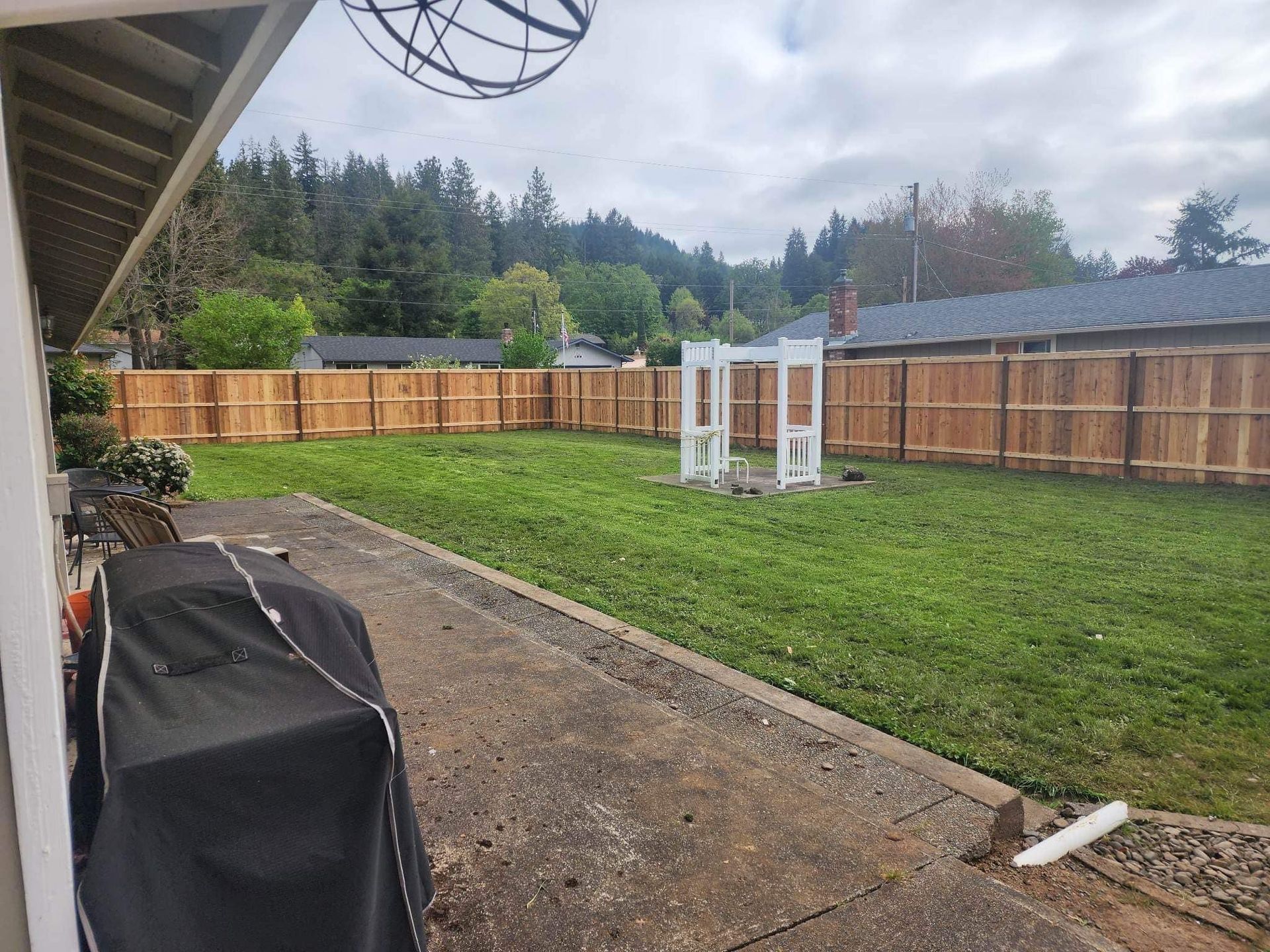 Backyard with green lawn, fenced perimeter, concrete patio, and a white swing set under cloudy skies
