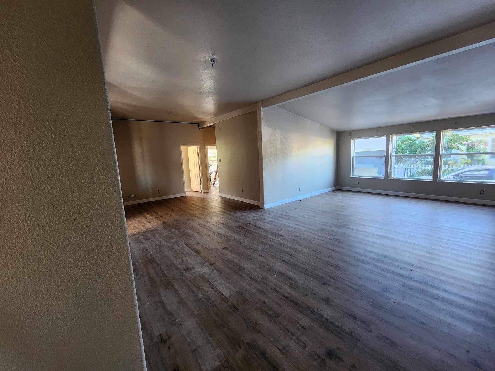 Empty living room with dark wood floors, beige walls, and large windows letting in daylight.