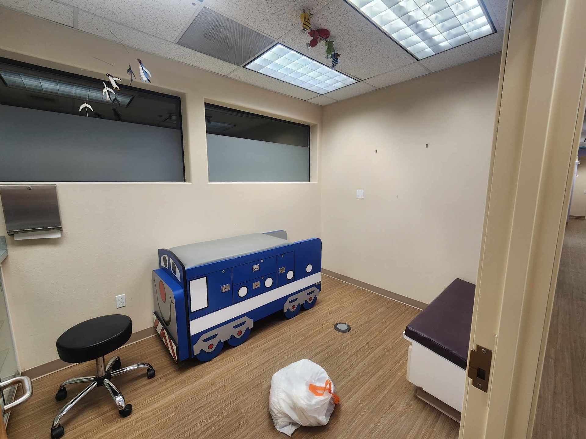 Small clinical room with a blue exam table, stool, wall sink, and fluorescent ceiling lights.