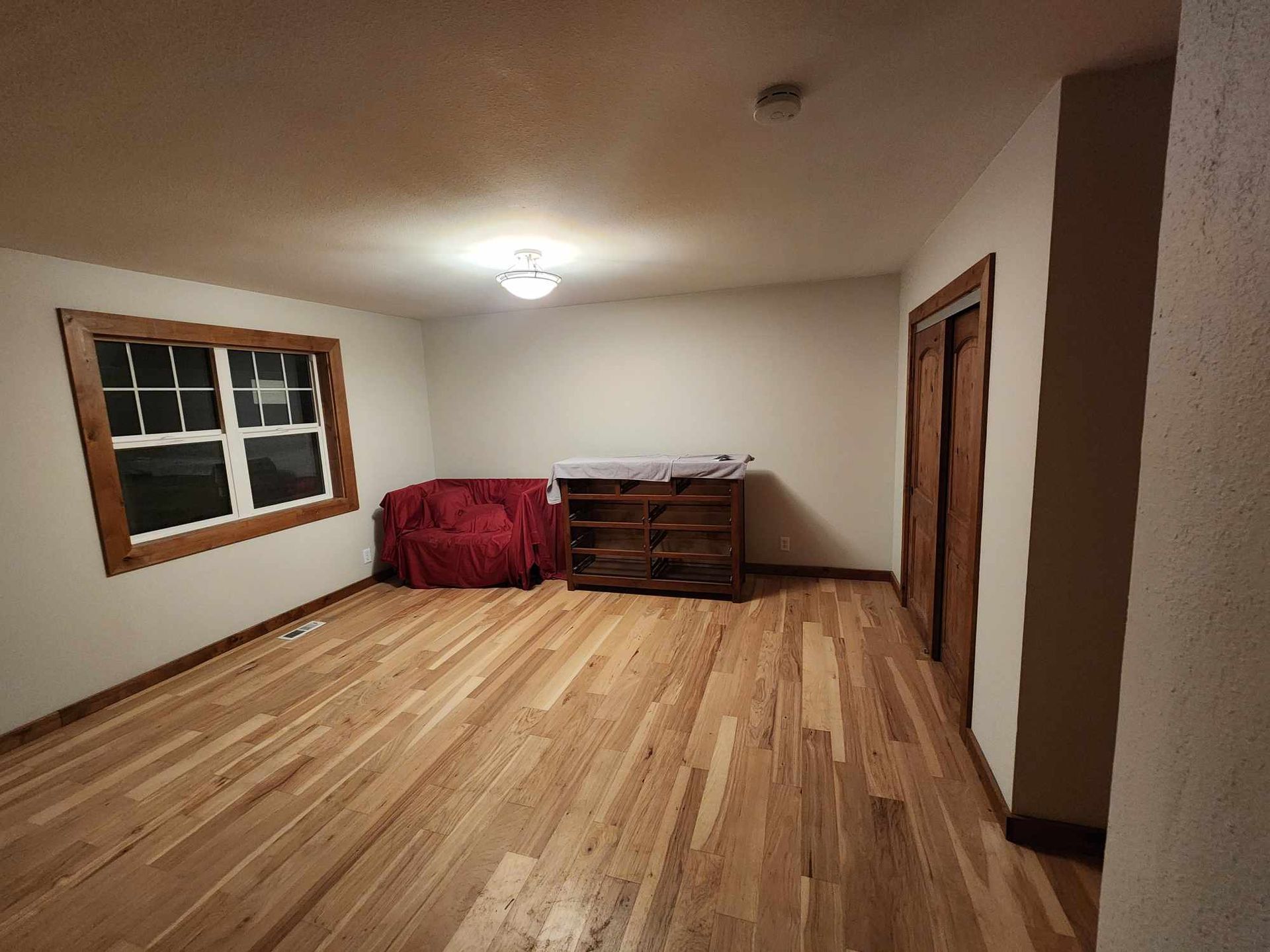 Empty unfurnished room with hardwood floors, window, overhead light, and a red-covered dresser against the wall