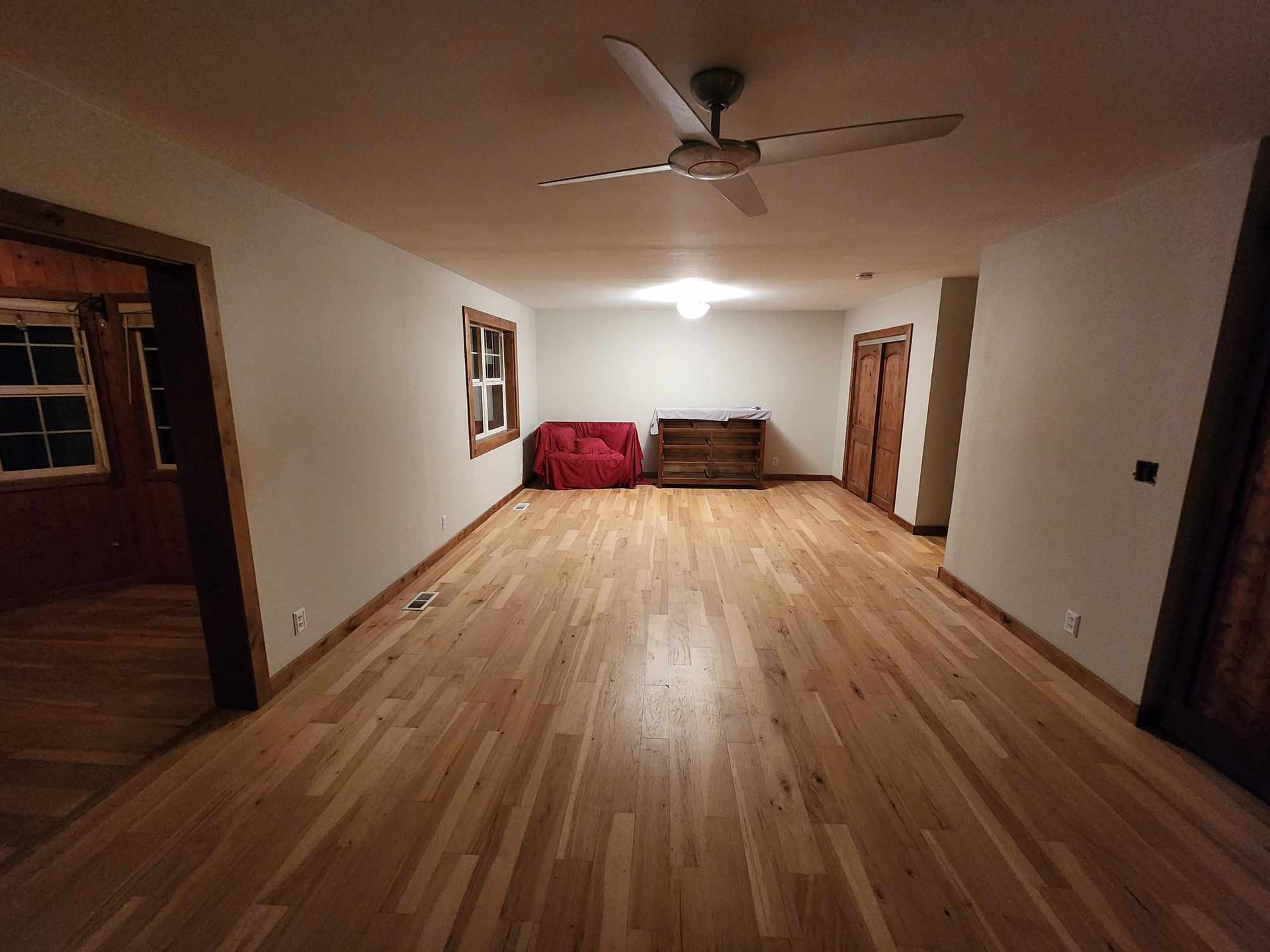 Empty living room with hardwood floors, ceiling fan, white walls, and red seating against the back wall.