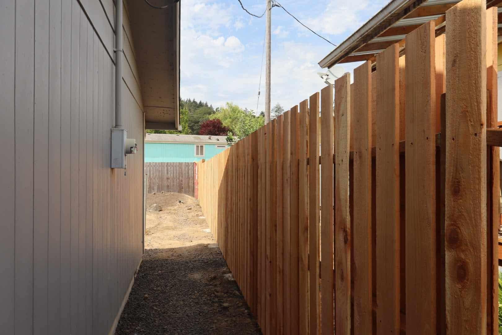 Narrow outdoor passage between a gray house wall and a tall wooden fence, leading to a backyard gate.