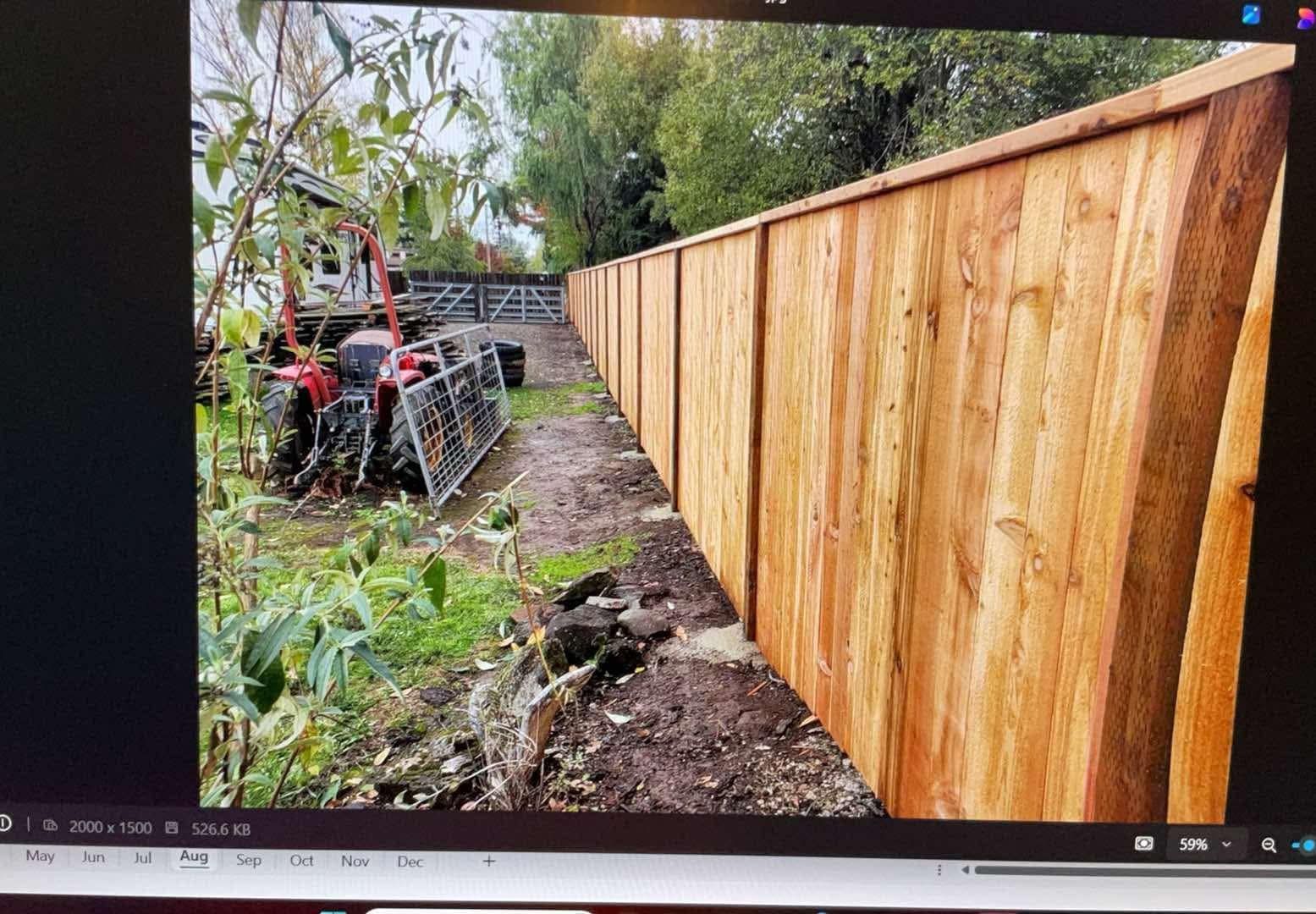 New wooden fence along muddy yard with a tractor on the left and trees overhead