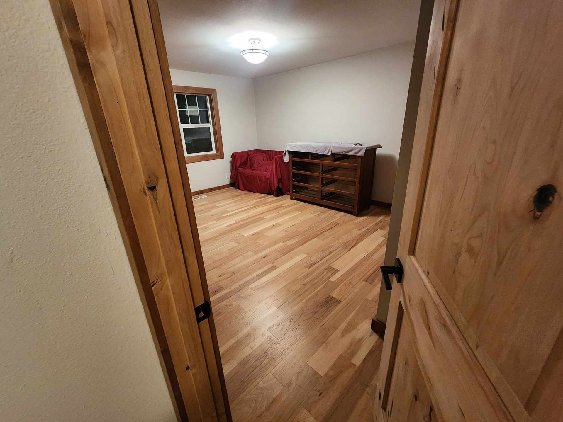 Open wooden doorway into a small bedroom with wood floors, a dresser, window, and ceiling light.