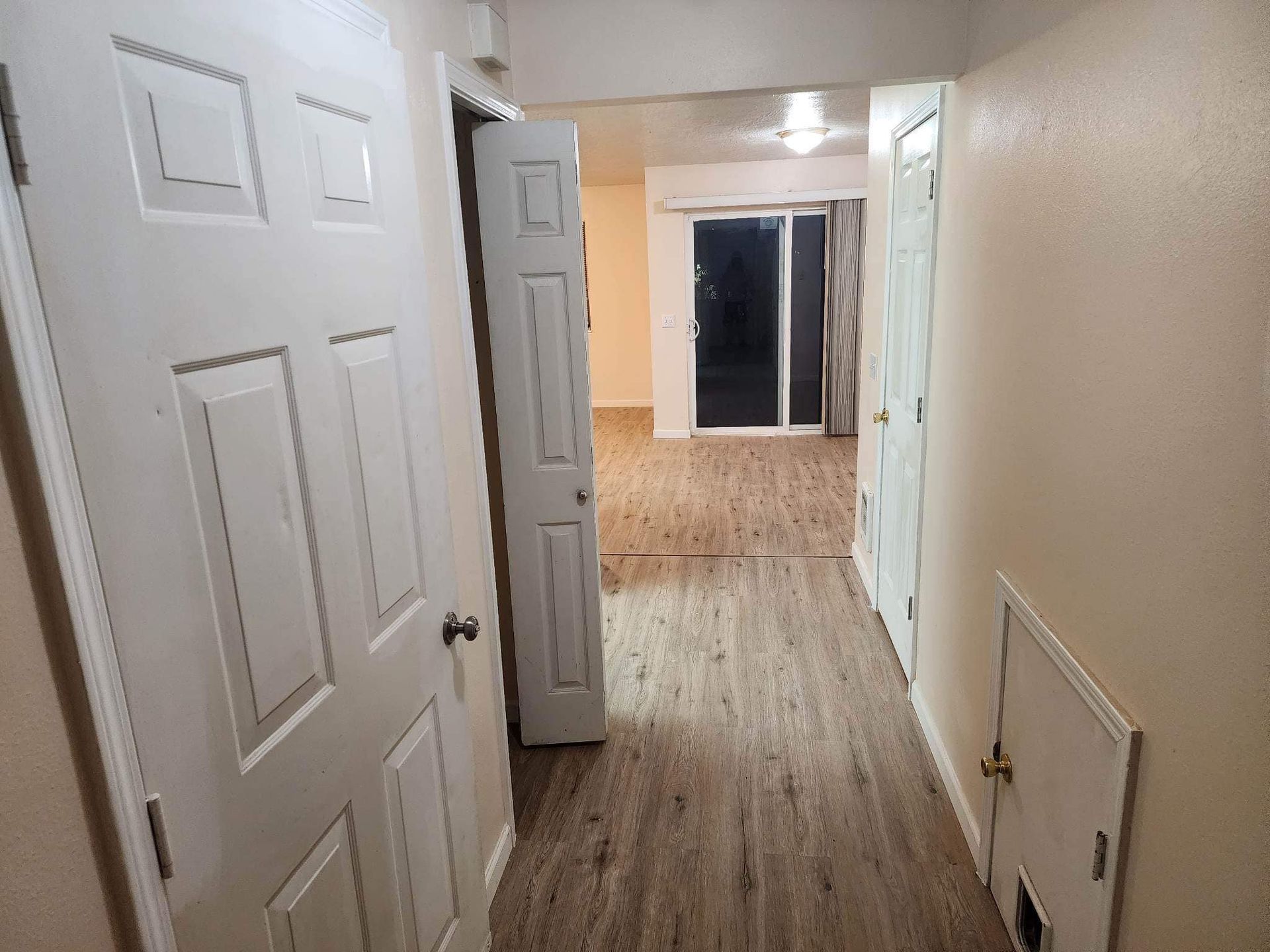 Empty hallway with open white doors, beige walls, and wood-look flooring leading to a darker room.