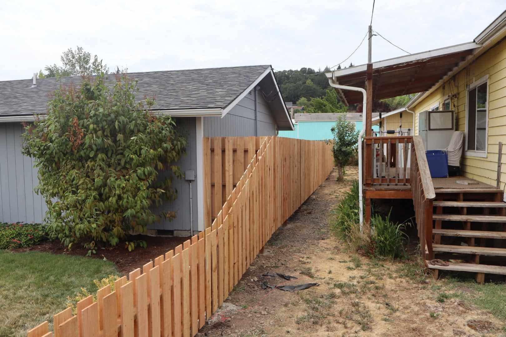 Backyard with wooden fence between a gray shed and a yellow house porch with steps