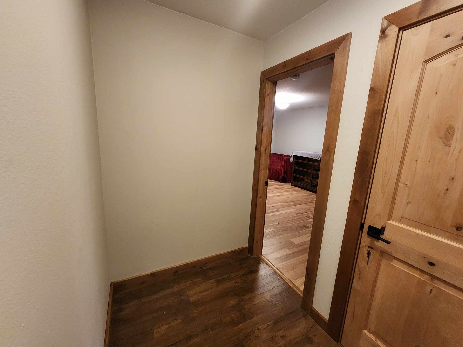 Empty hallway with wooden floor and trim, beige walls, and an open doorway to a room with a red couch.