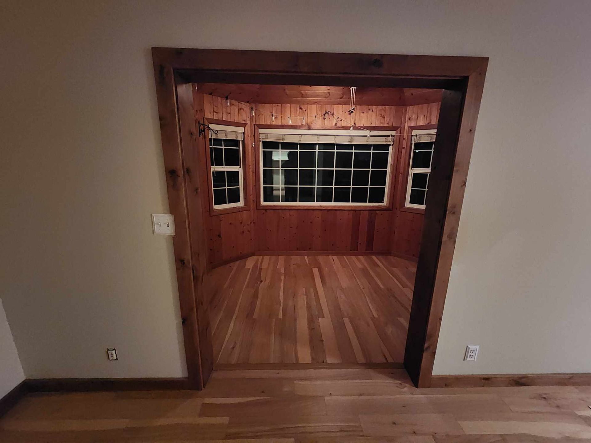 Wood-paneled room framed by a wide doorway, with a large window and hardwood floor inside.