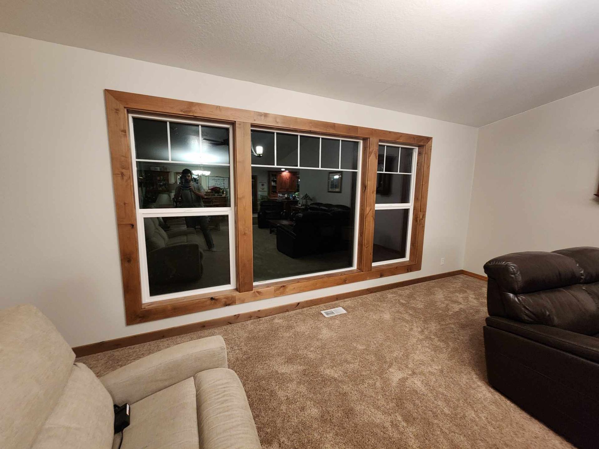 Empty living room with large wood-trimmed windows and beige carpet, partial view of sofa and armchair.