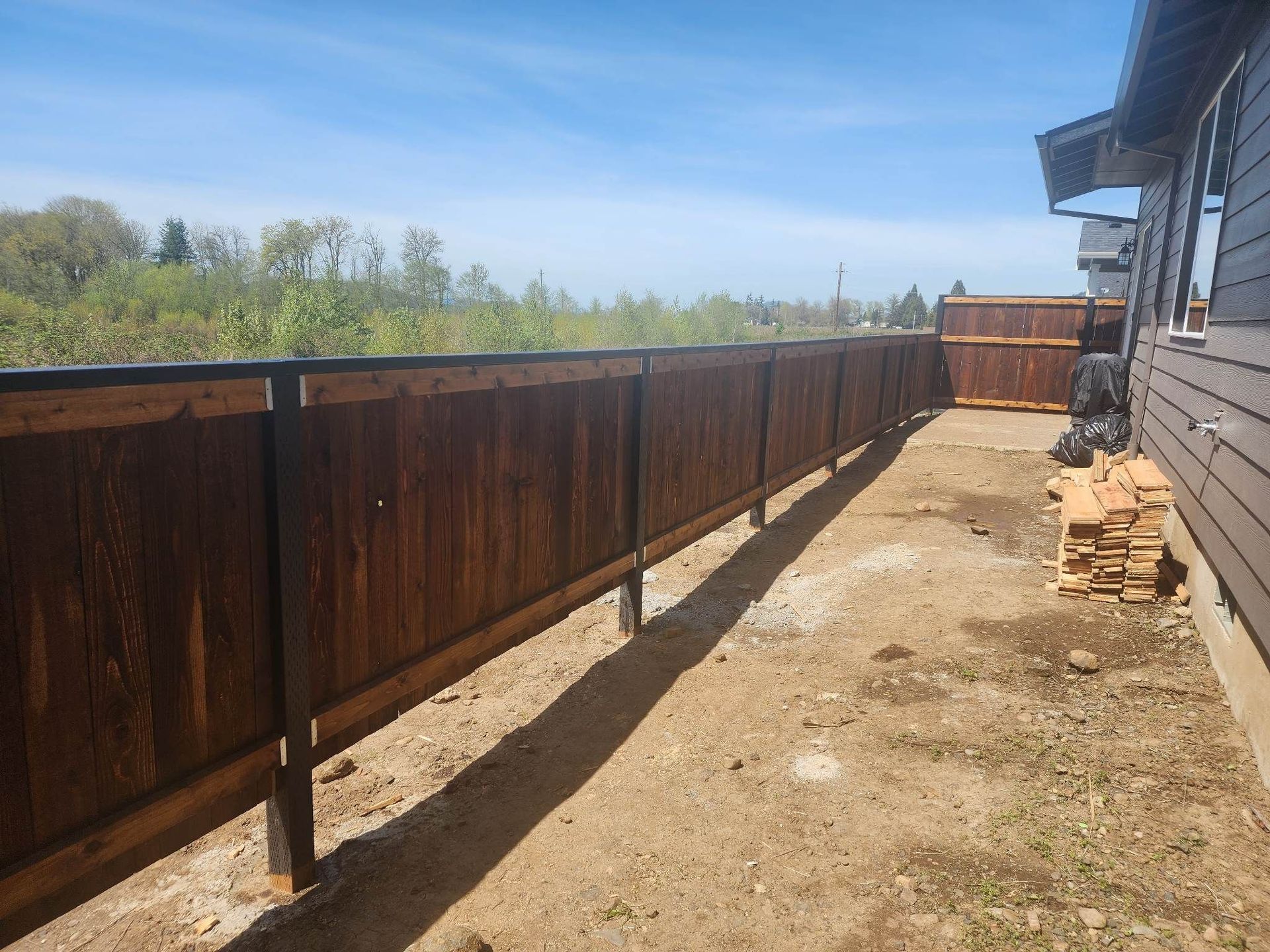 Rust-colored wooden fence along a narrow dirt path beside a house, under a clear blue sky.