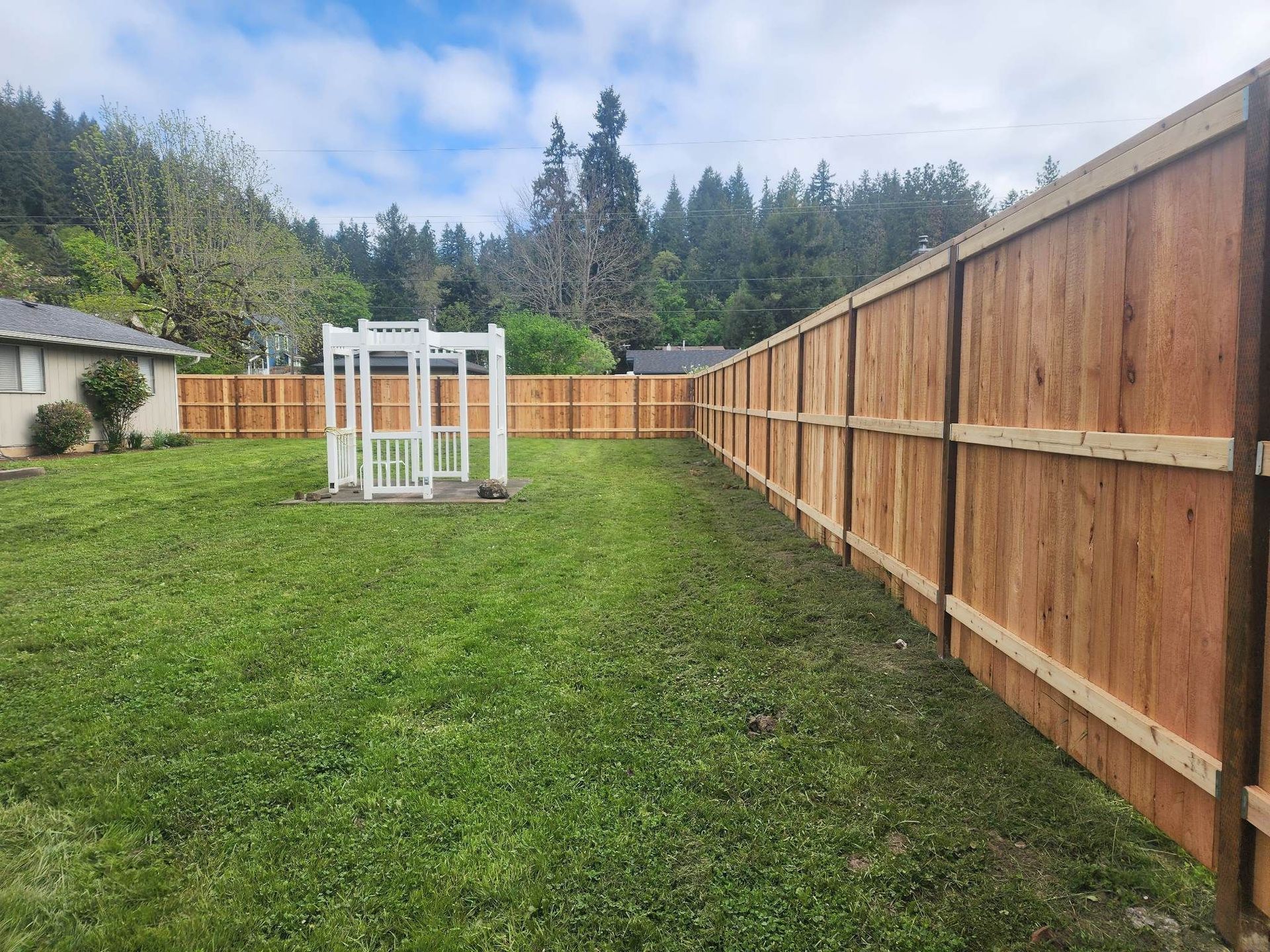 Backyard with green lawn, wooden fence, and small white greenhouse under cloudy sky