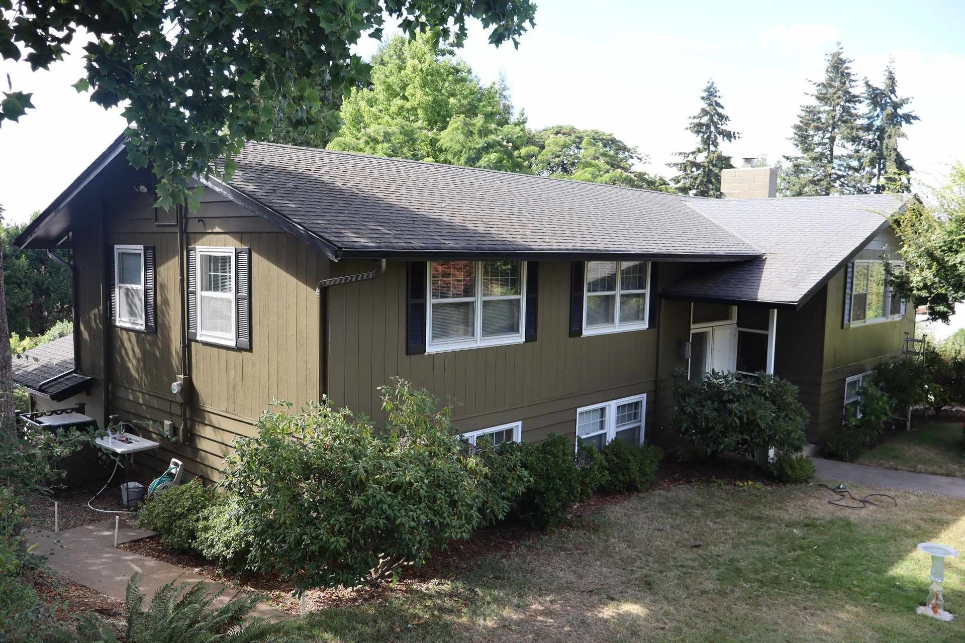 Small tan house with a dark roof, bushes in front, and trees in the background.