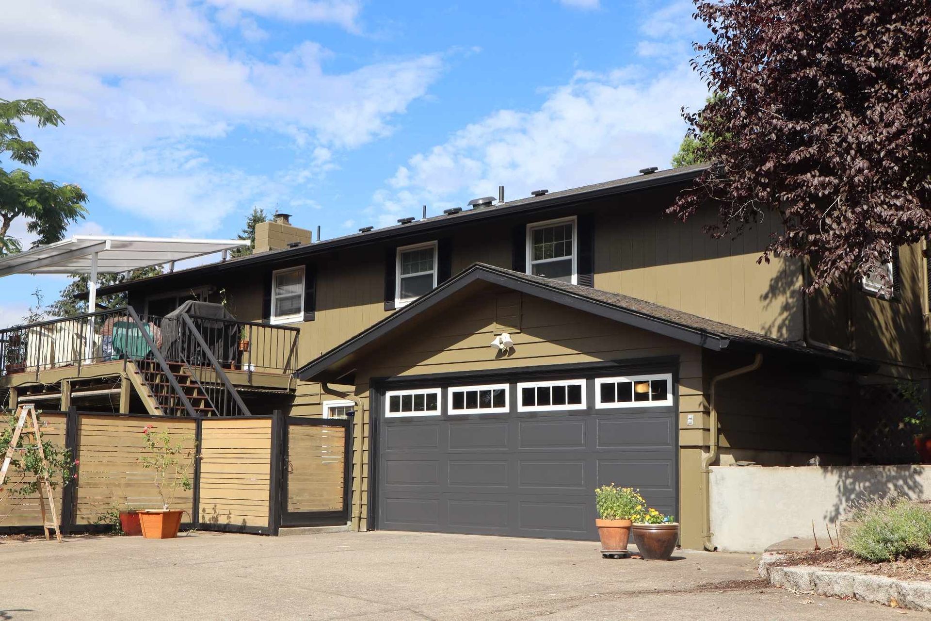 Suburban house with tan siding, dark garage door, and wooden fence under a blue sky